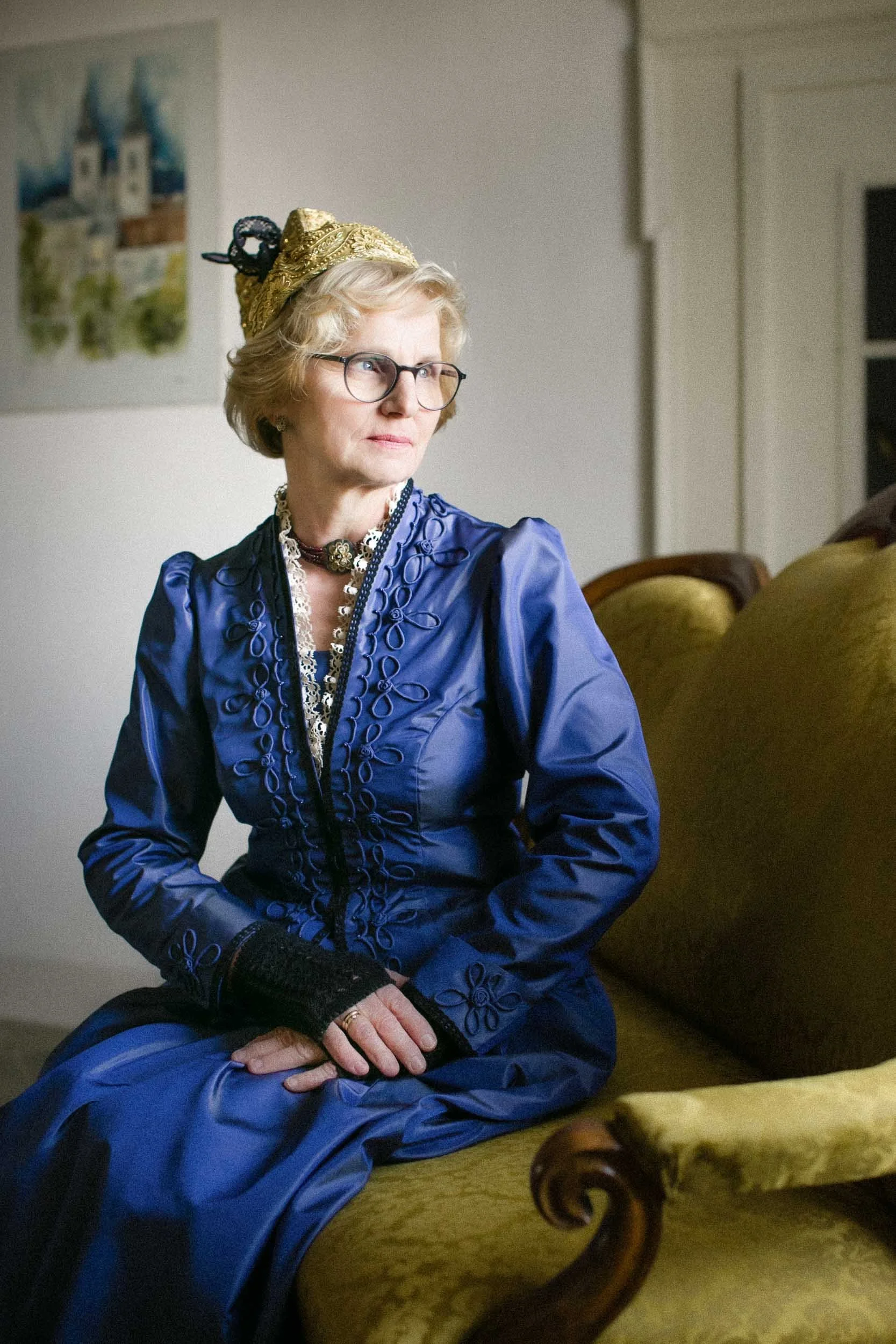 Eine ältere Frau mit Brille trägt ein elegantes, blaues Kleid mit Stickerei, sitzt auf einem grünen Sofa und schaut nach rechts.