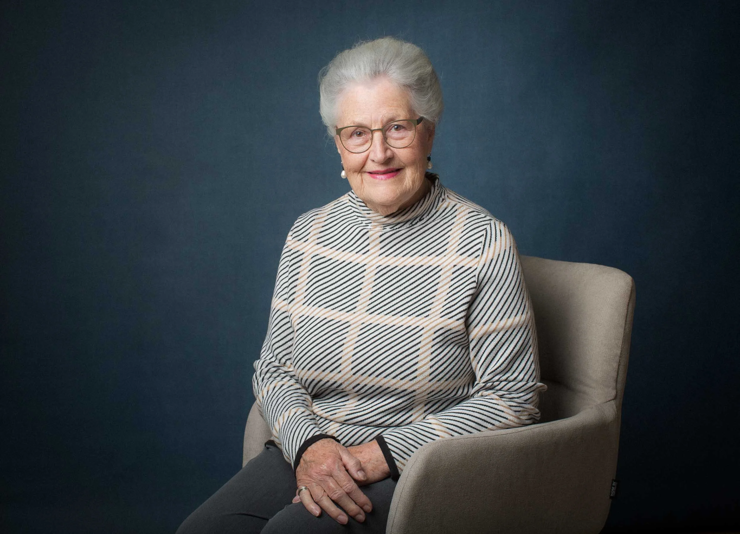 Ältere Frau mit Brille sitzt auf einem grauen Stuhl vor einem dunklen Hintergrund und lächelt.