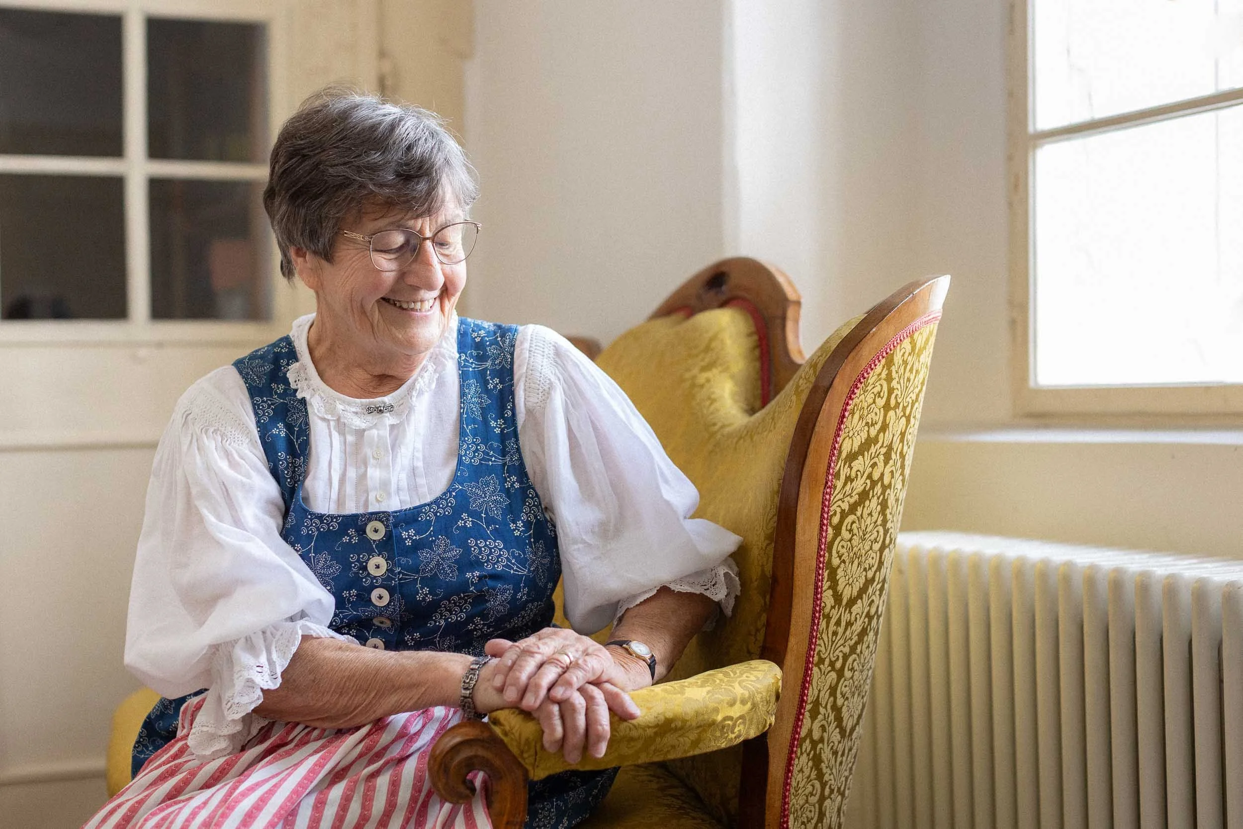 Eine ältere Frau mit Brille, die auf einem antiken gelben Sessel sitzt, lächelnd.