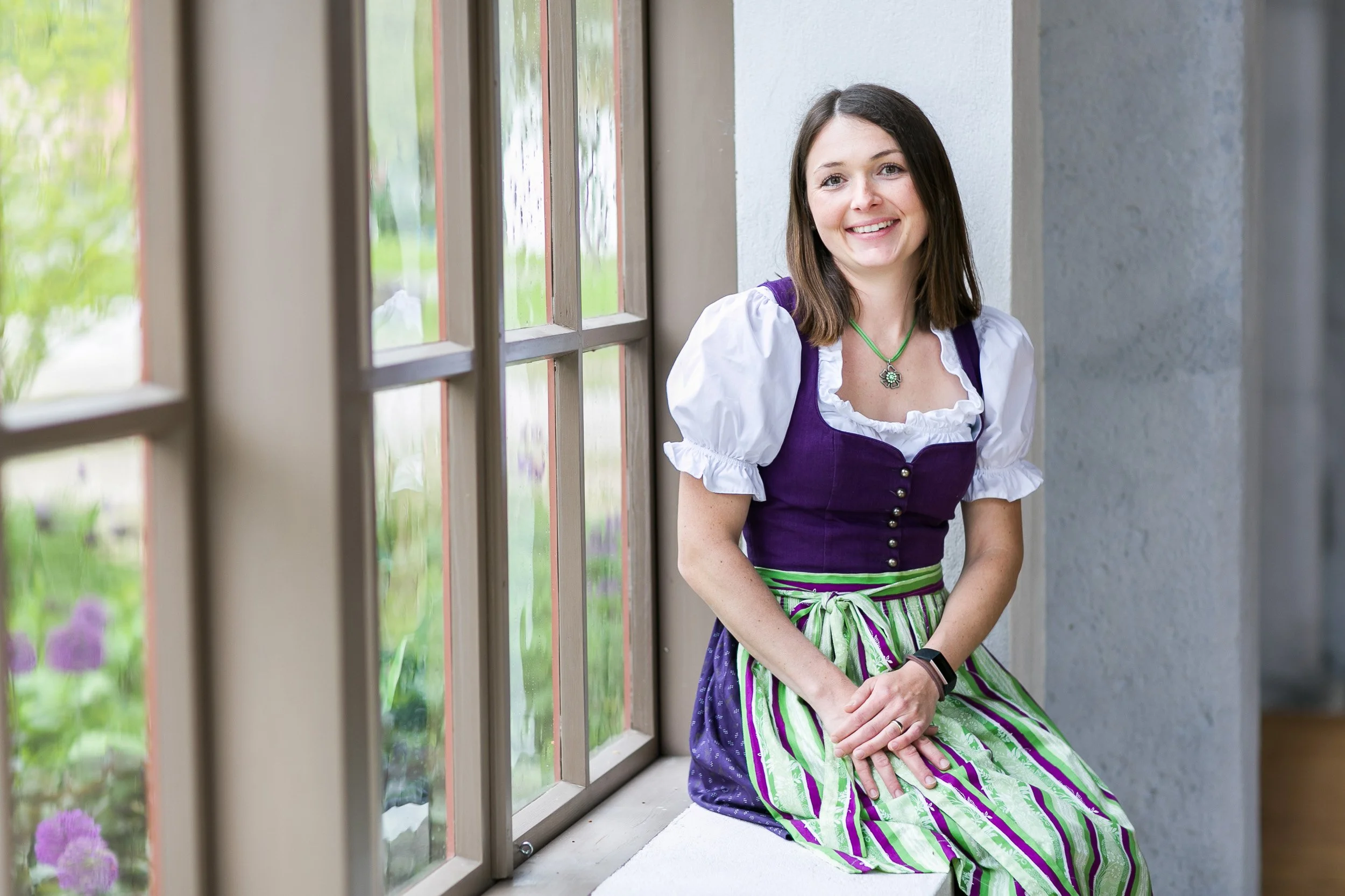 Frau in traditioneller bayerischer Tracht sitzt neben einem Fenster, lächelt und schaut nach vorne.
