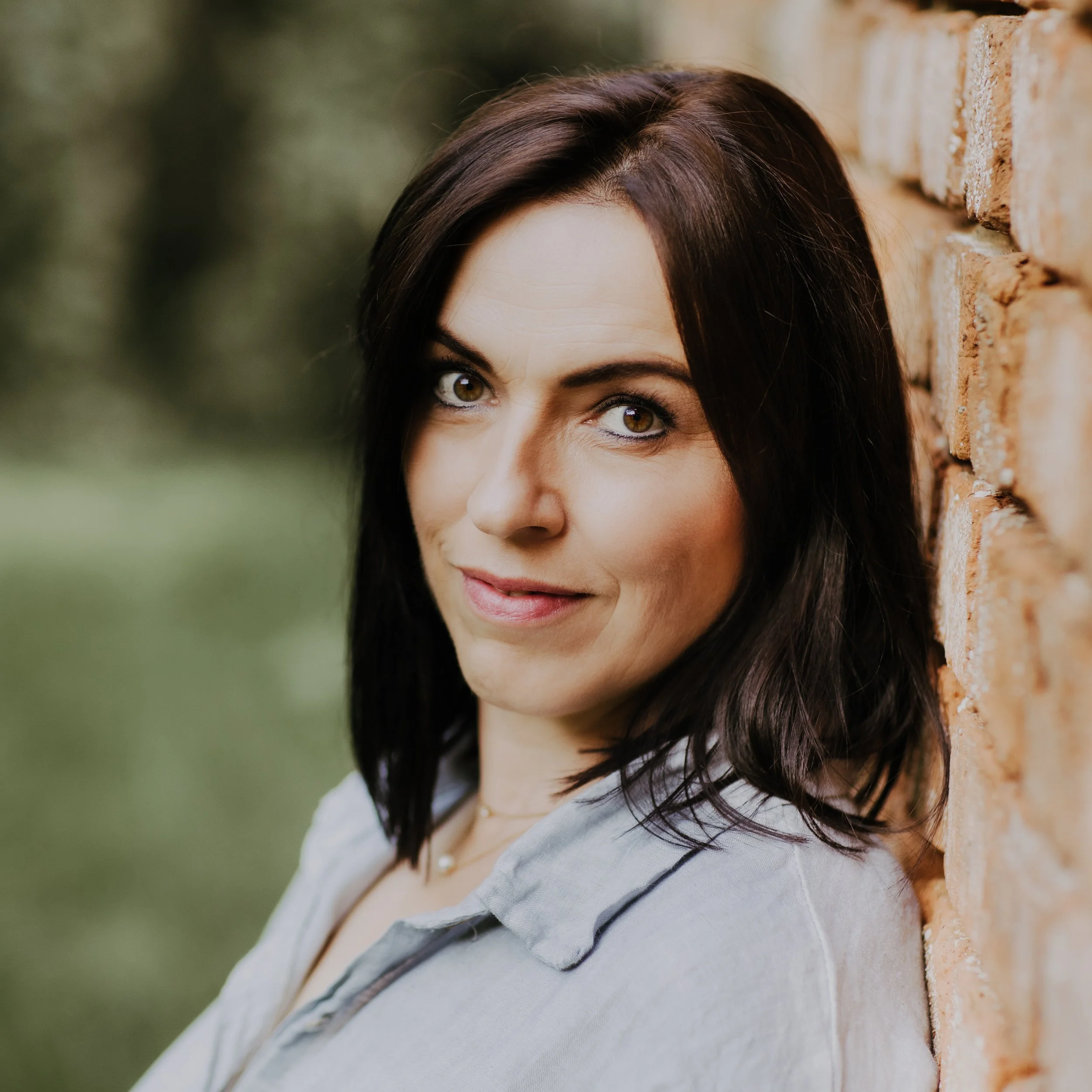 Nahaufnahme einer Frau mit schwarzen Haaren, die an einer Ziegelmauer lehnt, mit einem grünen, unscharfen Hintergrund.