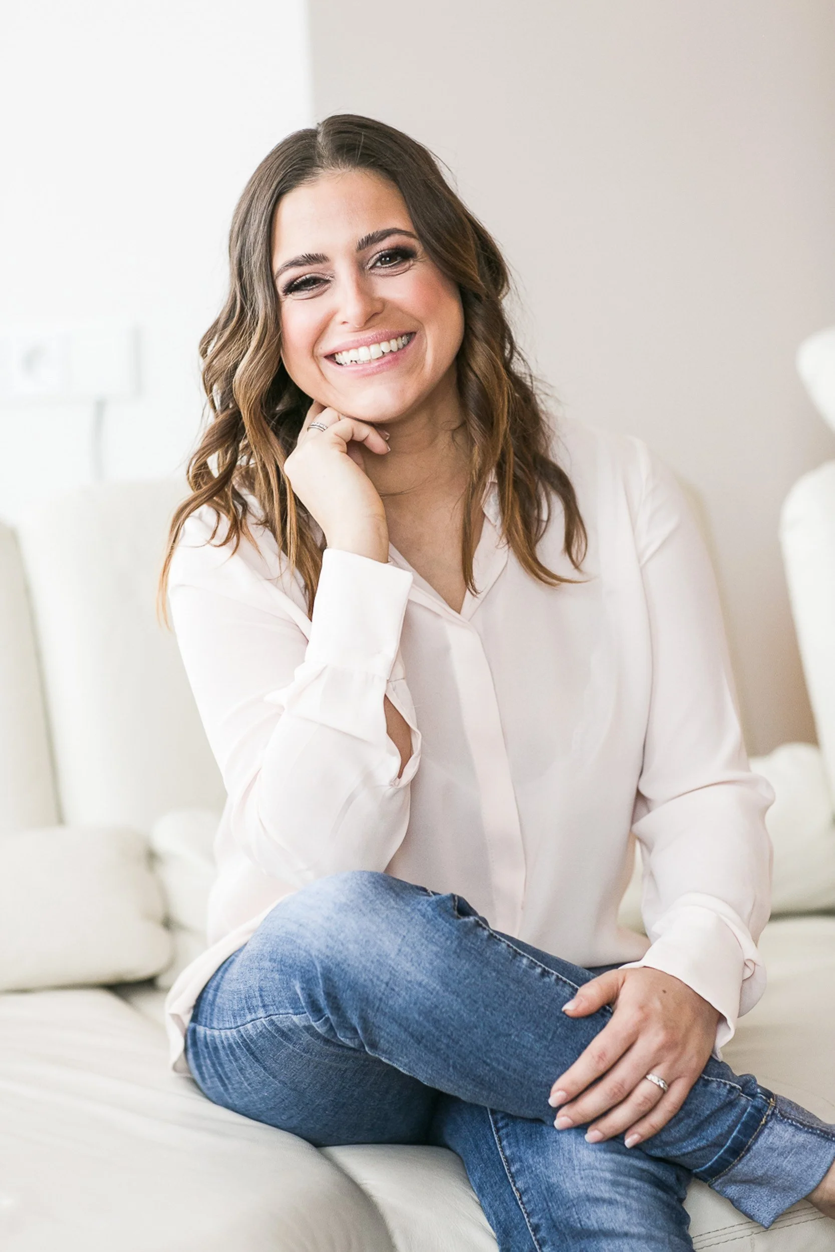 Eine lachende Frau mit braunen, lockigen Haaren, casual Kleidung, sitzend auf einem weißen Sofa in einem hellen Raum.