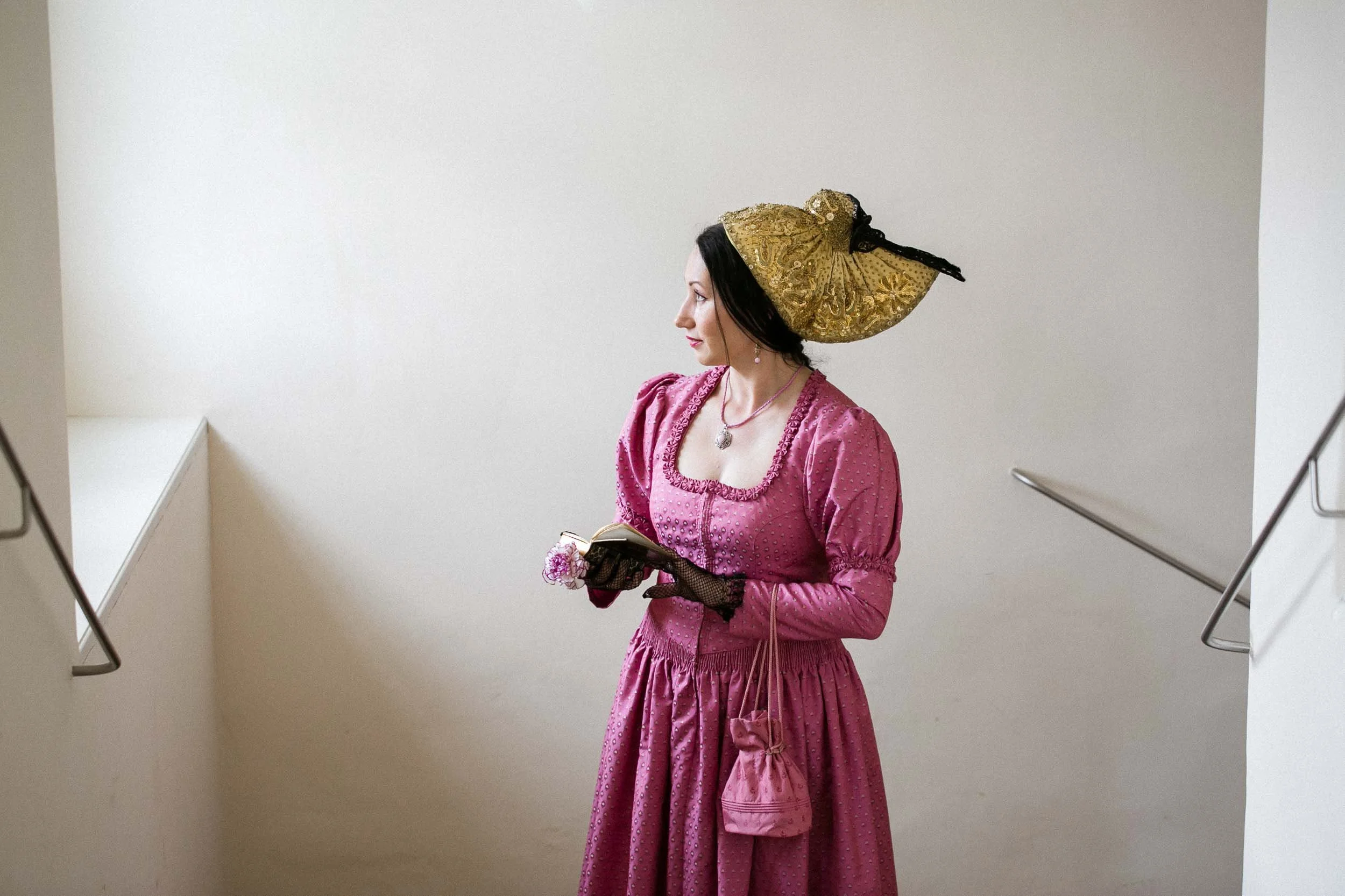 Frau in 18. Jahrhundert Kleidung mit goldener Haube, pinkem Kleid, schwarzen Handschuhen, liest ein Buch im Treppenhaus.