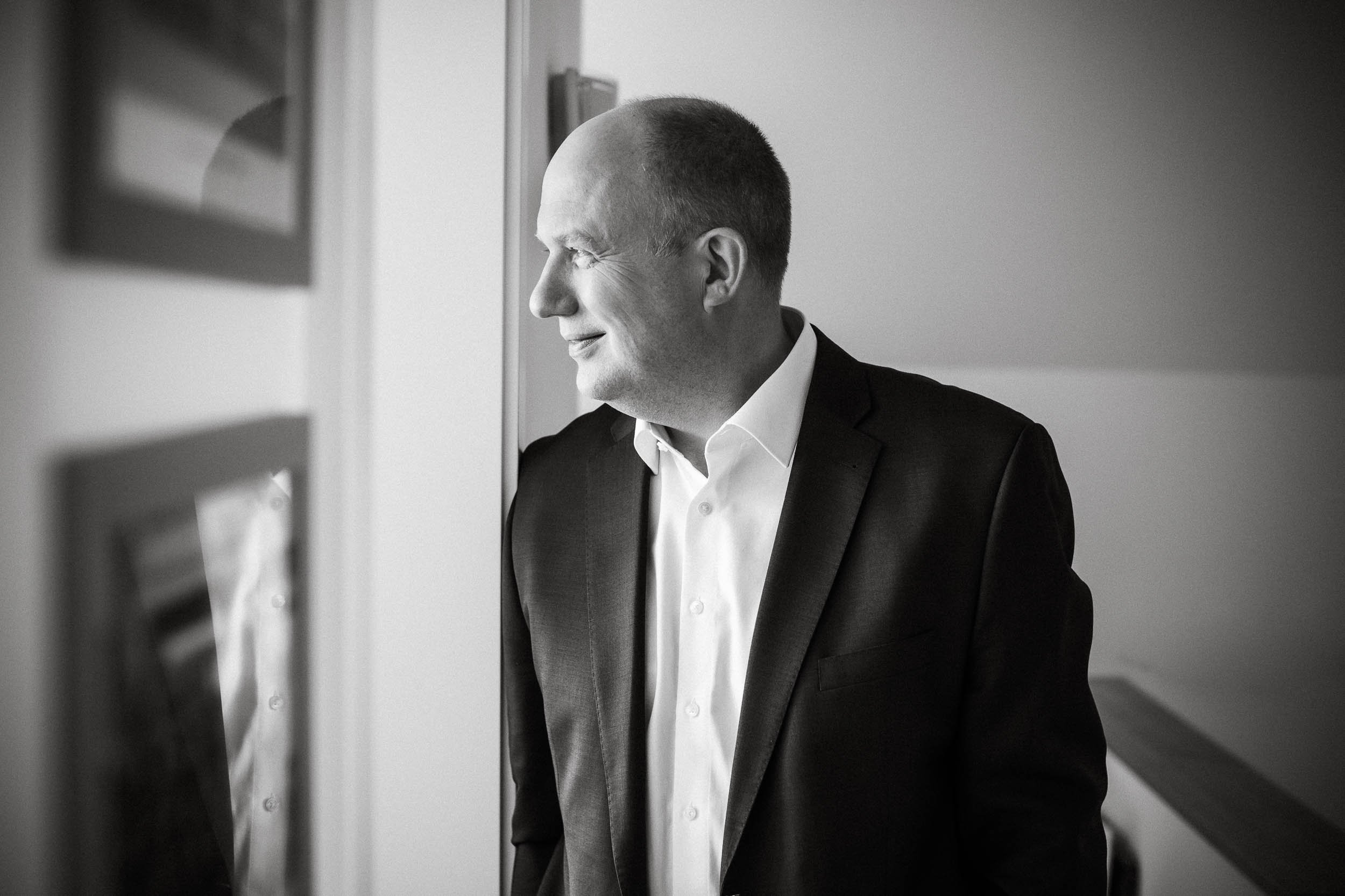 Mann in schwarzem Anzug und weißem Hemd schaut nach draußen, während er an einer Wand steht.