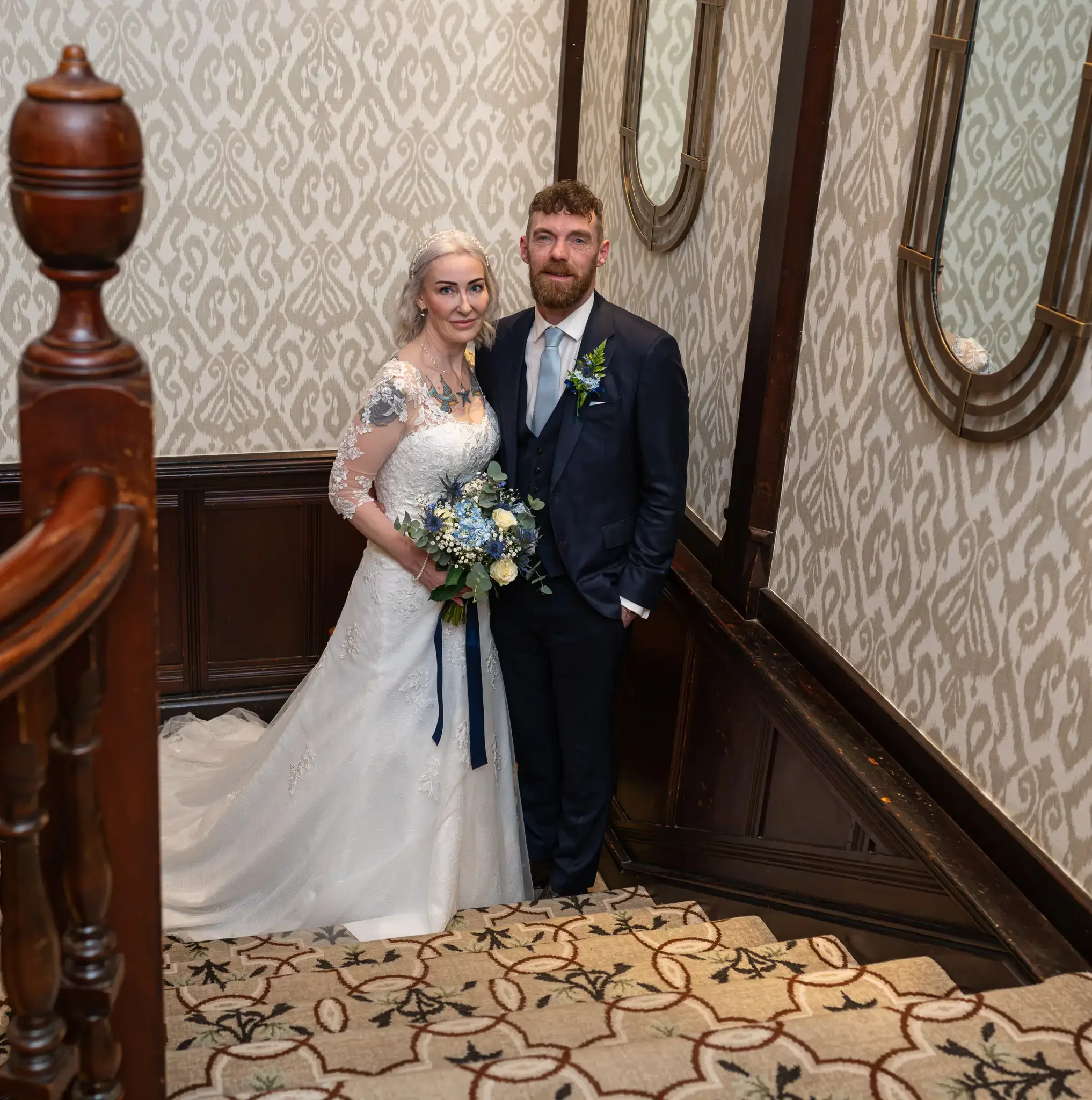 An indoor wedding portrait showcasing the grand, historic interior of the Piersland House Hotel in Troon. The composition uses the dark wood ceiling beams and ornate banisters of the upper hallway to create strong leading lines that draw the eye thro