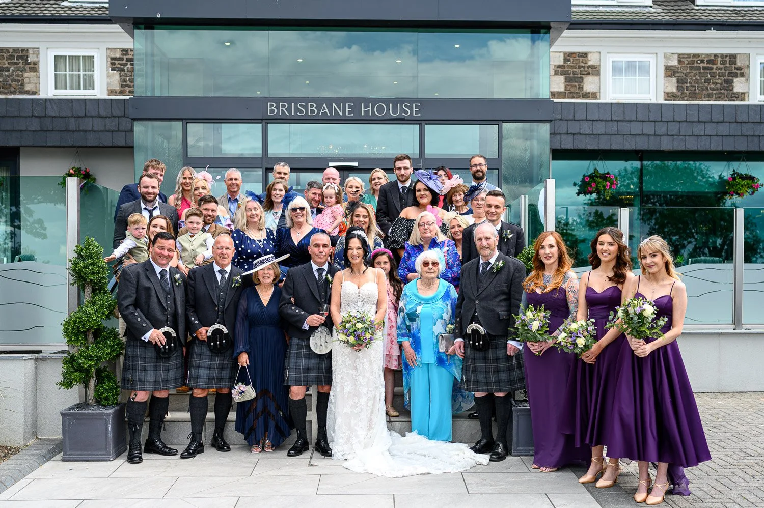 A large group of wedding guests gathered in front of the modern glass entrance of Brisbane House Hotel.