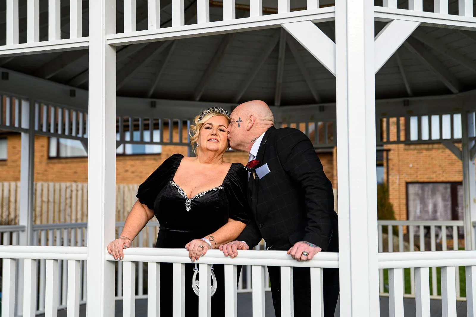 A couple stands on a white gazebo, with the man kissing the woman's cheek; the woman wears a black dress and a tiara, wedding-style, and the man is dressed in a dark suit with tattoos visible on his head and hands.