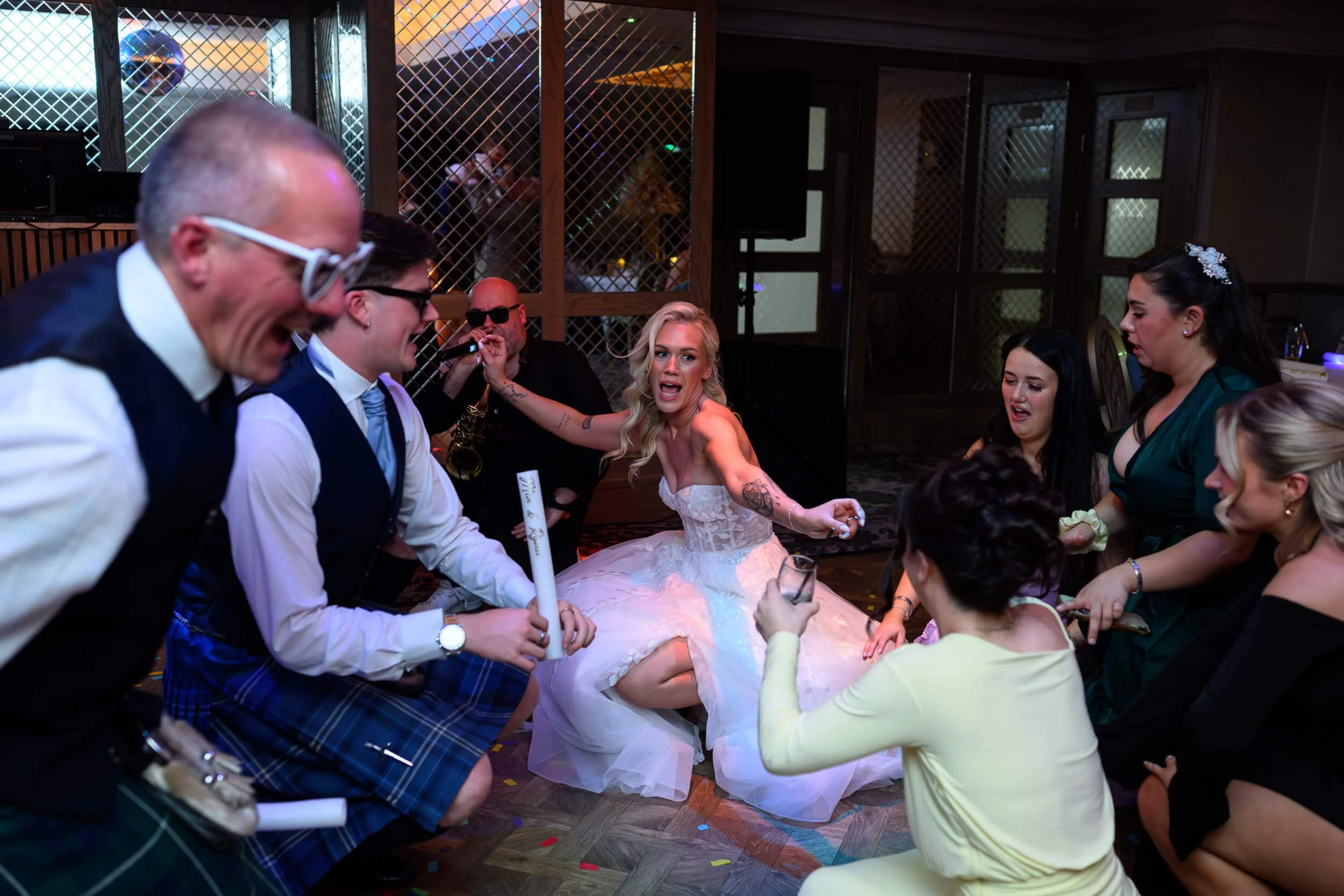 High-energy wedding reception moment at Brisbane House Hotel in Largs, with the bride kneeling on the dancefloor surrounded by cheering guests. Glow sticks, live music, and joyful expressions capture the electric party atmosphere and the unforgettabl