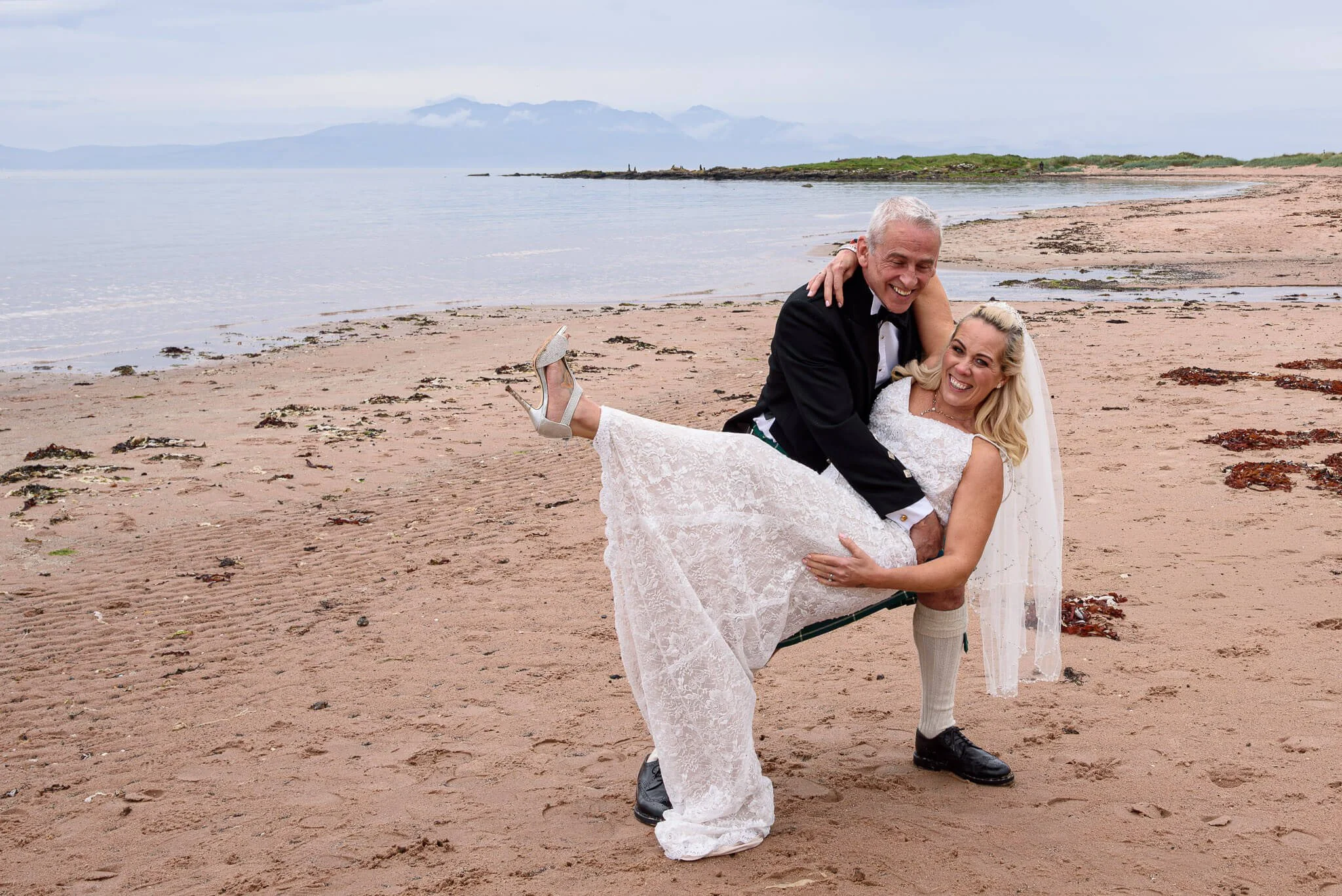 A groom in a traditional Scottish kilt lifting his laughing bride into the air on Seamill Beach, Ayrshire, with the sea and the hills of Arran in the background.