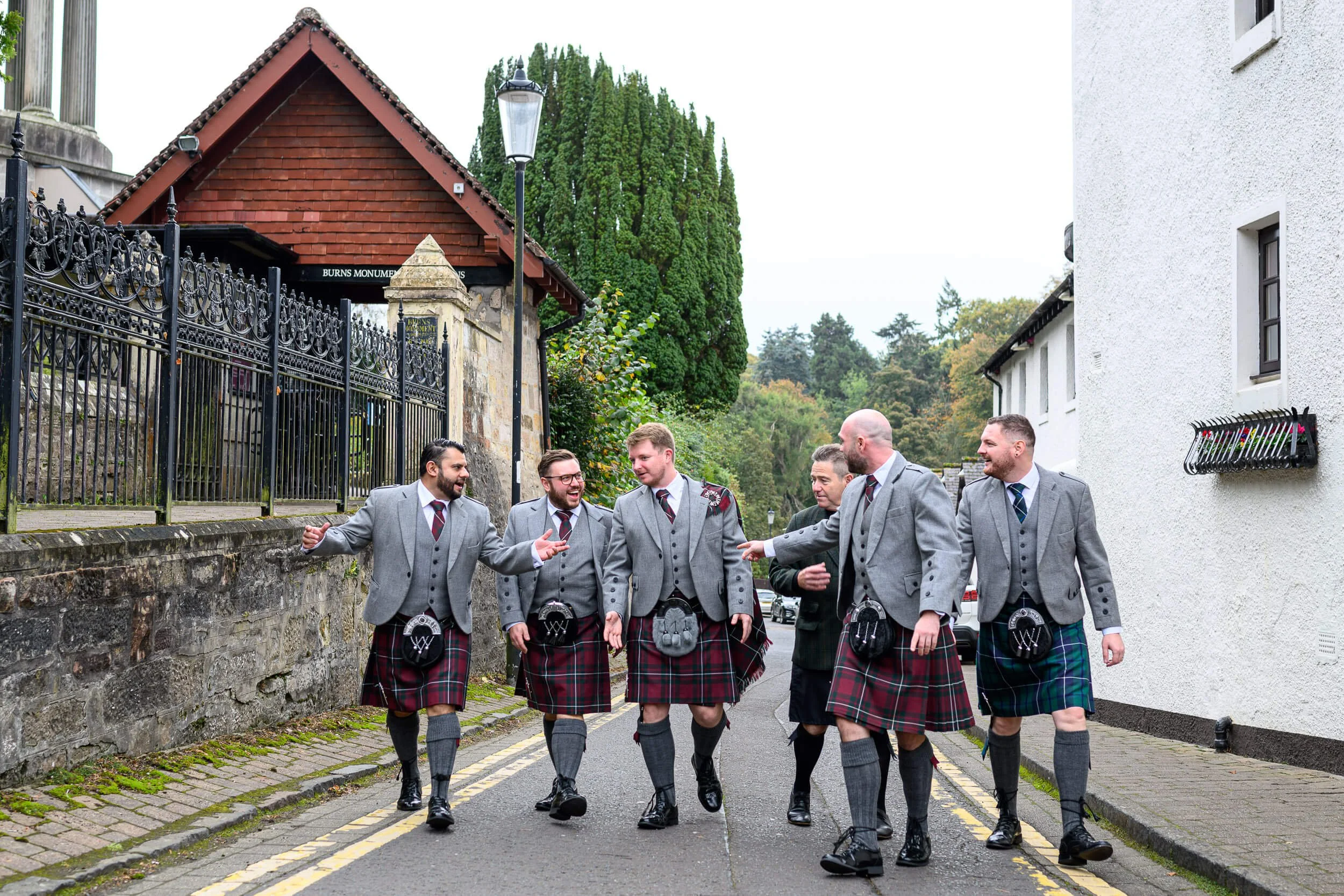A stylish group of groomsmen walk confidently down the cobbled street beside Brig O’ Doon in Alloway, dressed in traditional Scottish kilts and grey tweed jackets. Captured in a relaxed, candid moment before the ceremony, the image showcases camarade