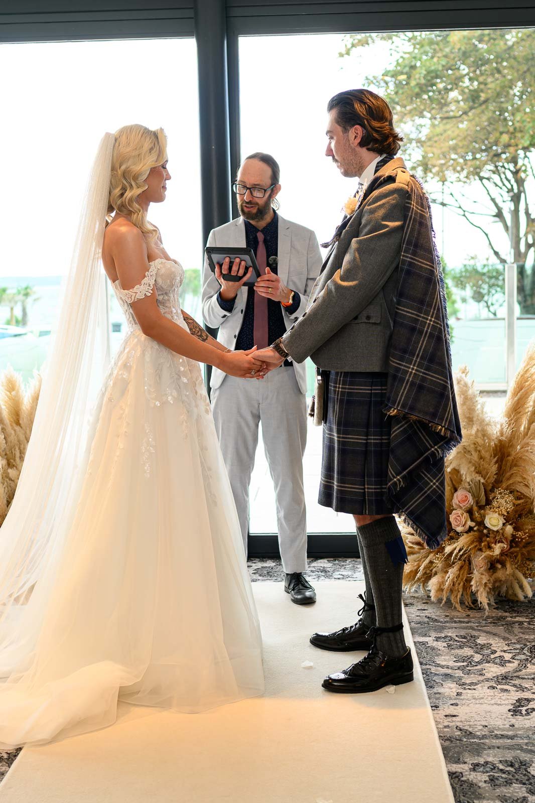 A couple getting married during a wedding ceremony, holding hands and exchanging vows, with a celebrant officiating. The bride has long blonde hair and is wearing a white wedding gown, while the groom is dressed in a traditional kilt with a grey jacket.