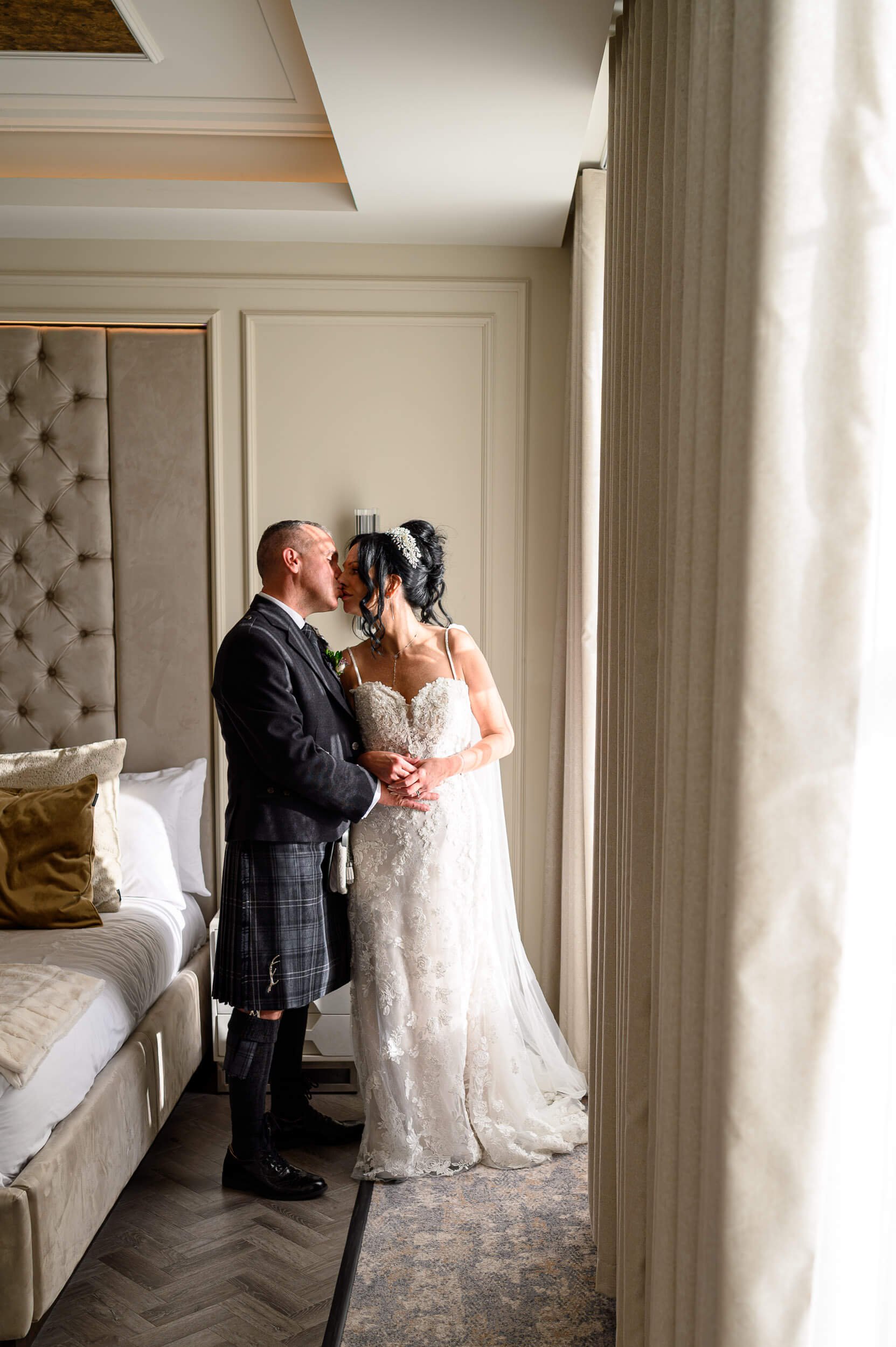 A romantic window-light portrait at Brisbane House Hotel in Largs. Soft natural light pours through the curtains as the bride and groom share a quiet kiss in their hotel suite, creating an intimate and timeless wedding moment.