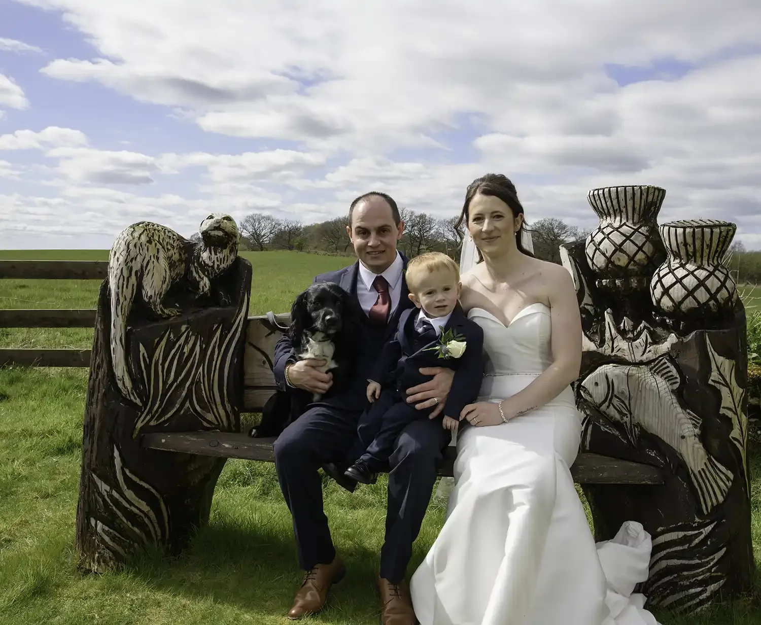 Bride Samantha and groom Daniel sitting with their child and dog on a carved wooden bench at The Gathering at Woodhead Farm in Chryston on their wedding day