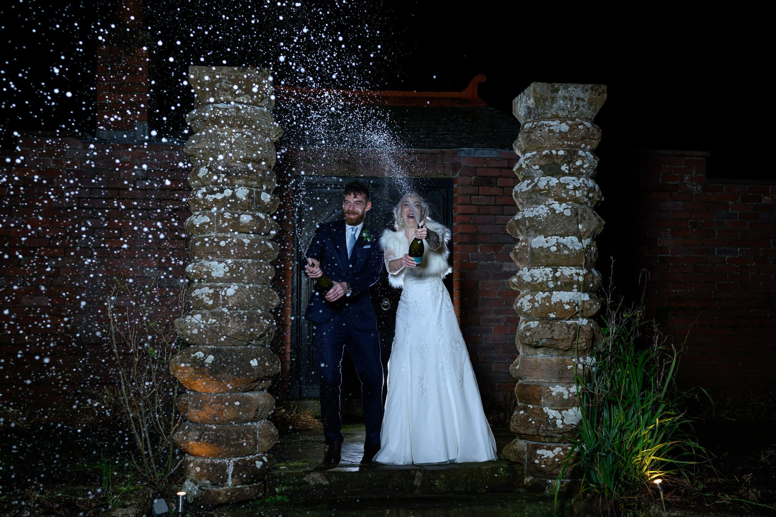 A fun and energetic night-time wedding portrait at Piersland House Hotel in Troon. The newlyweds celebrate in style beneath the stone archway with a champagne spray moment, beautifully lit against the dark Ayrshire sky. Captured using off-camera flas