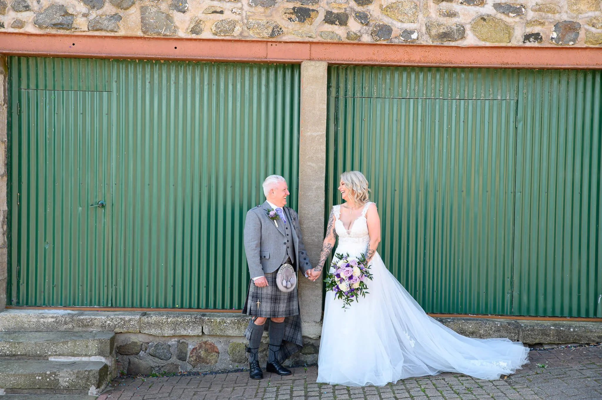 A bride in a flowing white gown and a groom in a grey kilt laughing together against a backdrop of green corrugated metal and stone walls at Harelaw Farm in Ayrshire.