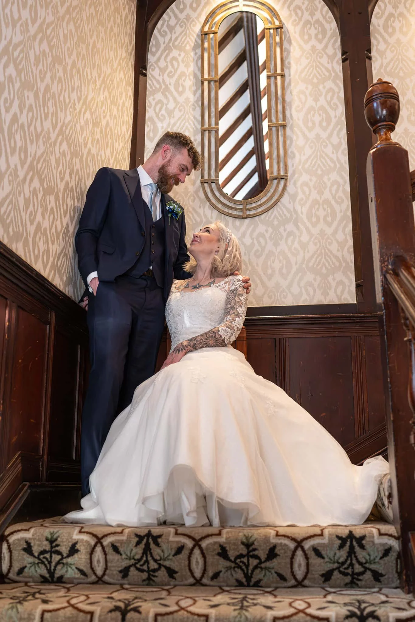 A striking indoor portrait captured within the historic Piersland House Hotel in Troon. The image utilizes the grand, dark-wood architectural beams and ornate banisters of the hallway to create deep leading lines and a sense of scale. The groom is po