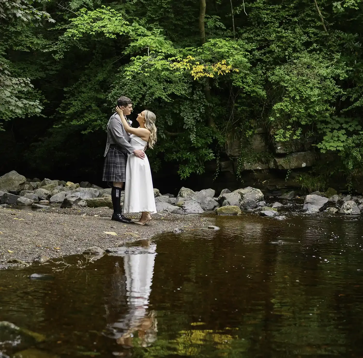 Bride and groom embracing by the river at Dean Castle Country Park in Kilmarnock