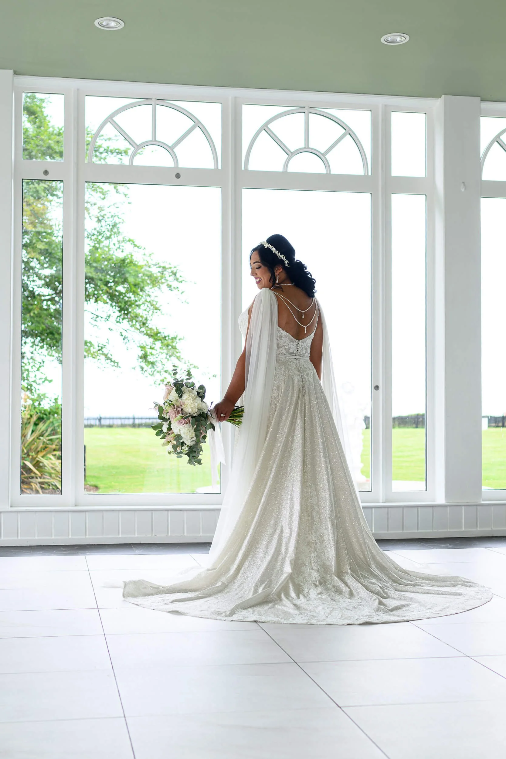 Elegant bridal portrait at Lochgreen House Hotel in Troon, captured in soft natural window light. The bride stands gracefully in front of tall white arched windows, her full gown flowing beautifully across the floor as she holds her bouquet and looks