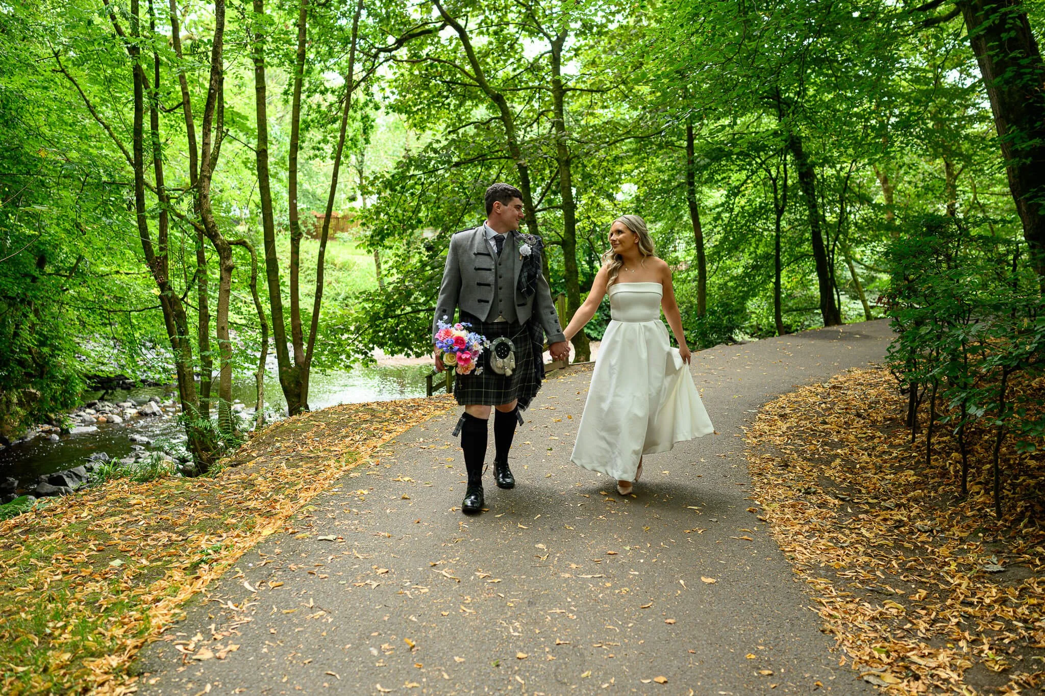 A bride in a strapless white gown and a groom in a grey kilt walking hand-in-hand along a leafy woodland path next to a stream at Dean Castle Country Park in Kilmarnock.