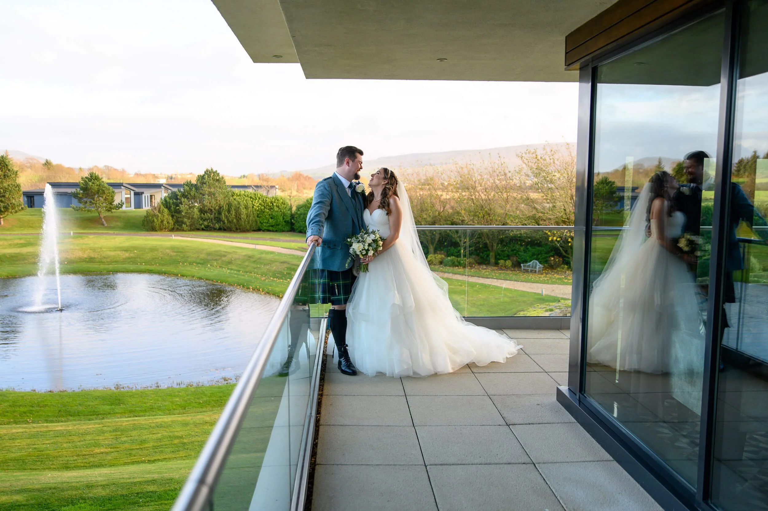 Romantic balcony portrait of the bride and groom at Lochside Hotel in New Cumnock, overlooking the fountain and Ayrshire countryside. Captured just before sunset, this natural moment shows the couple sharing a quiet laugh together as the bride’s gown