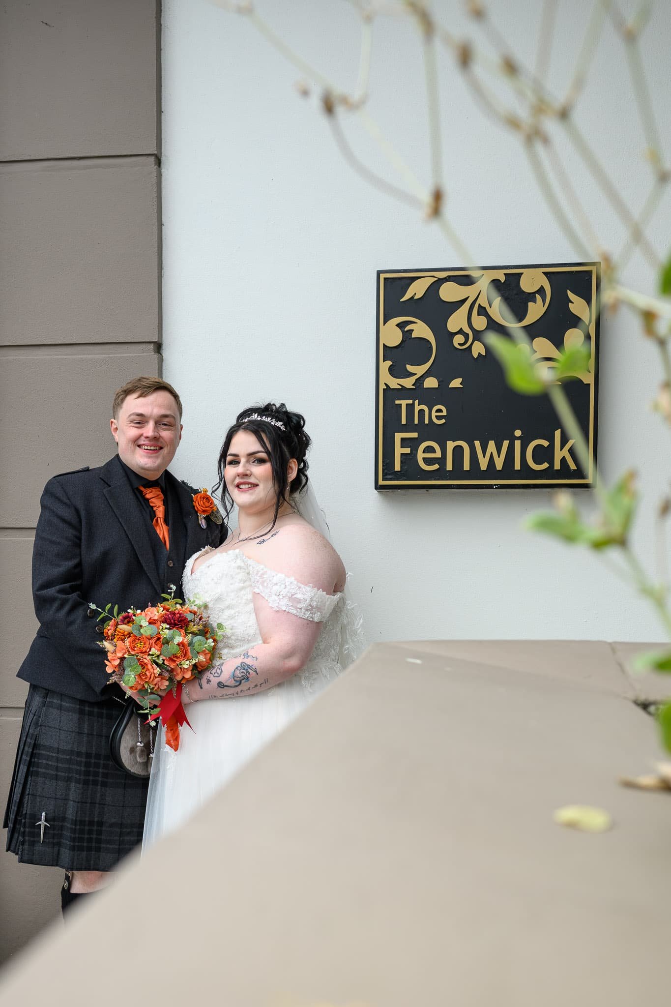 Bride and groom standing together outside The Fenwick Hotel in Ayrshire on their wedding day, smiling at the camera beside the venue sign while holding their bouquet.