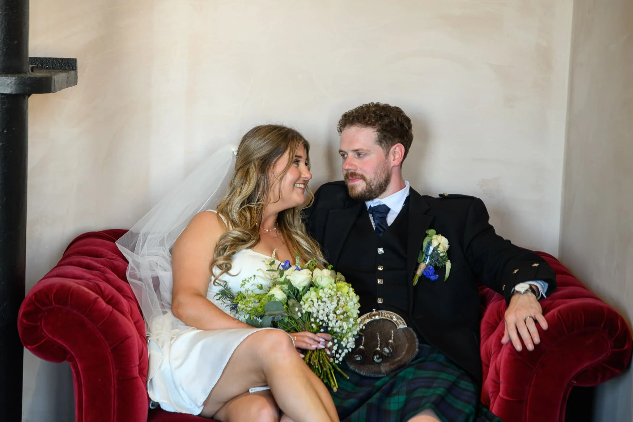 Bride and groom sitting together on a red sofa at The Engine Works in Glasgow, sharing a quiet moment and smiling at each other during their wedding day.