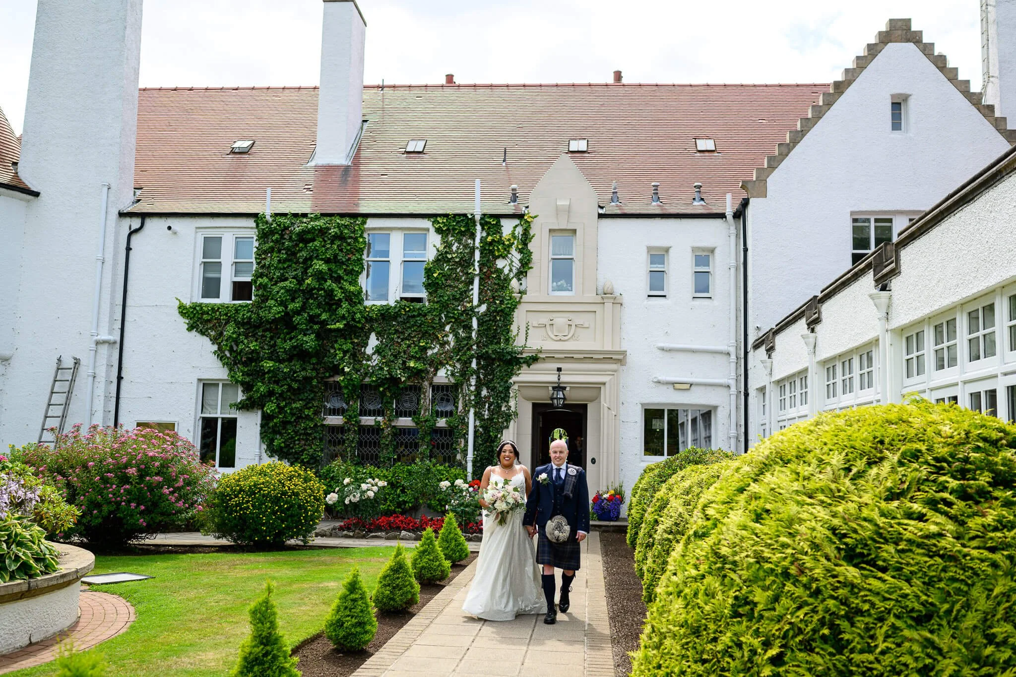 A bride and groom walking hand-in-hand down a stone path in the lush, ivy-covered gardens of Lochgreen House Hotel & Spa in Troon, Ayrshire