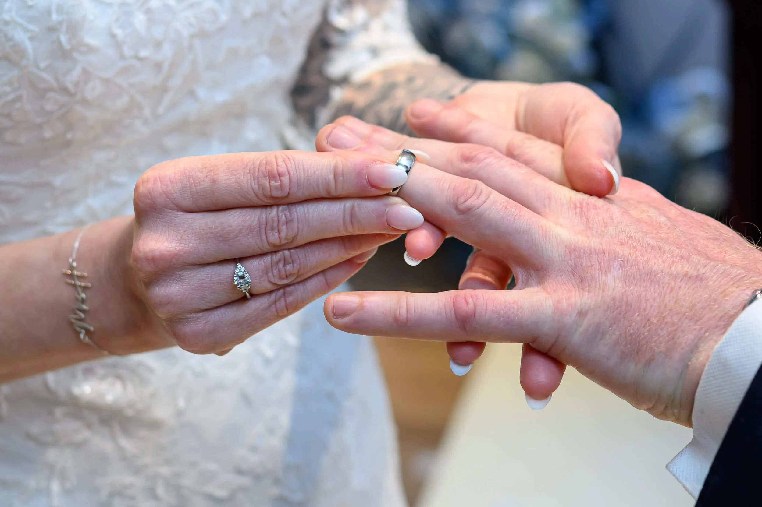 A close-up photograph of the wedding ring exchange at Piersland House Hotel in Troon. The bride gently places a silver wedding band onto the groom’s finger, with her engagement ring and manicured nails clearly visible. The shallow depth of field soft