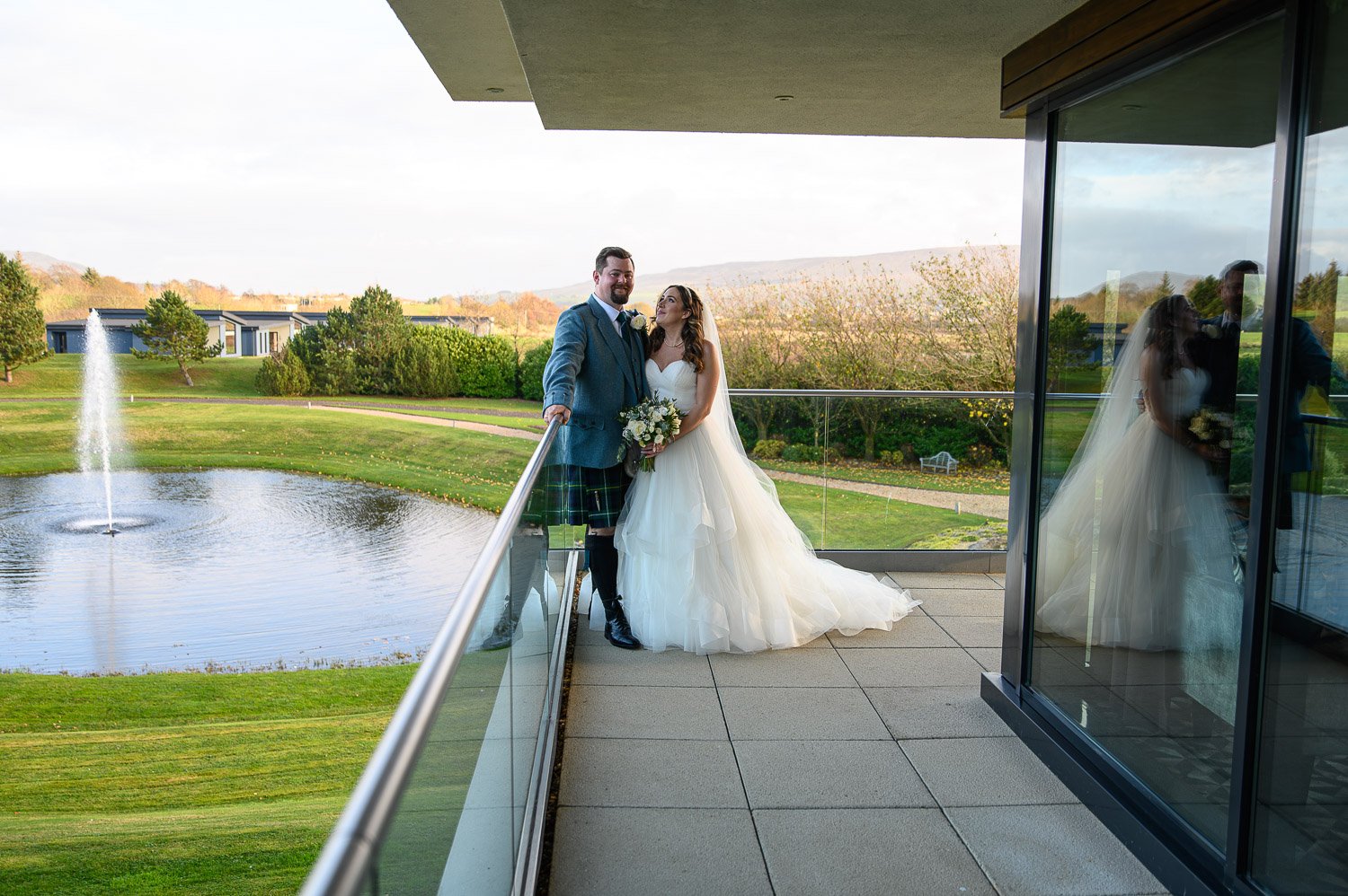 A bride and groom standing on the modern terrace balcony overlooking the fountain and hills at Lochside House Hotel.