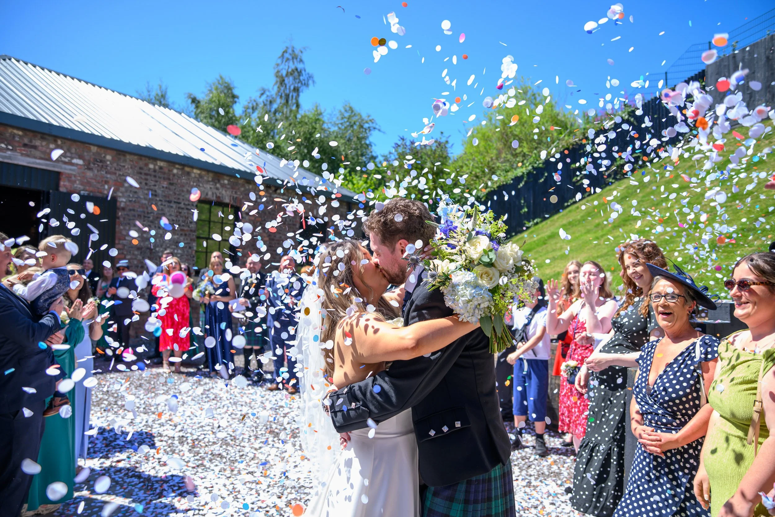 A joyful confetti kiss at Engine Works in Glasgow, captured as colourful confetti fills the air around the newlyweds. Guests cheer in the sunshine as the couple embrace, creating a vibrant, high-energy moment full of celebration and movement against 