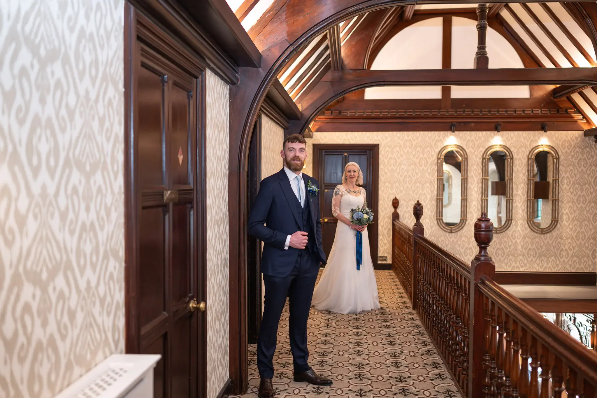 A wedding photo taken in a beautifully decorated indoor space at the Piersland House Hotel, which can be found in the town of Troon. The groom stands in the foreground on the left. The bride stands further down the hallway. The hotel interior, with i