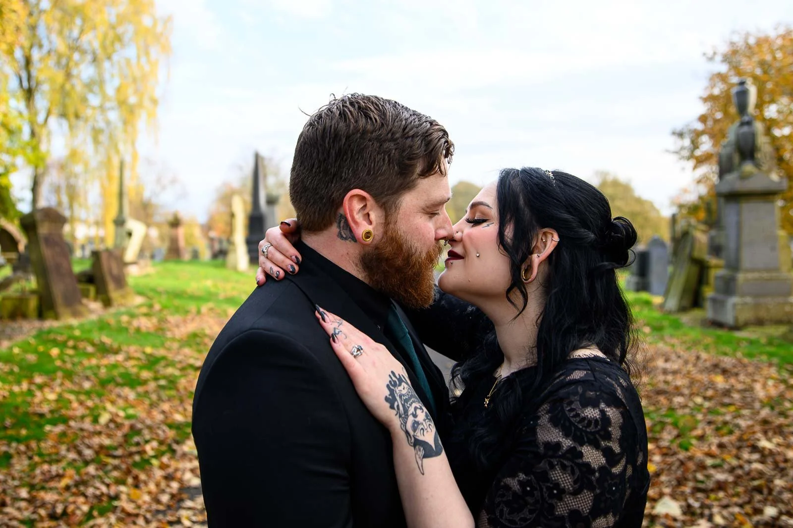 A couple with tattoos and piercings is embracing and about to kiss in a graveyard during autumn, with fallen leaves and tombstones in the background.