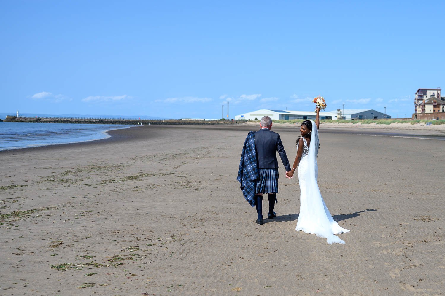 A relaxed coastal wedding portrait on Ayr beach, capturing a quiet moment as the bride and groom walk hand in hand along the shoreline. Natural, storytelling wedding photography in Ayrshire with the beautiful Scottish coastline as their backdrop.