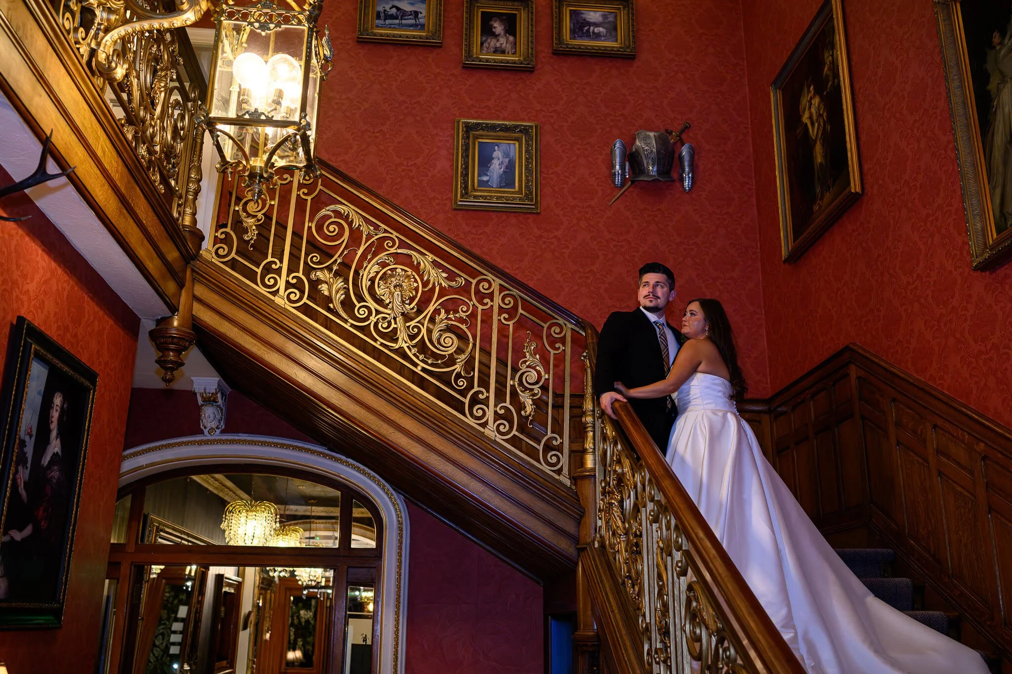 bride in a sleek white gown and a groom in a black suit standing on a magnificent, ornate gold and wood staircase at Auchen Castle, surrounded by red damask walls and historic portraits.