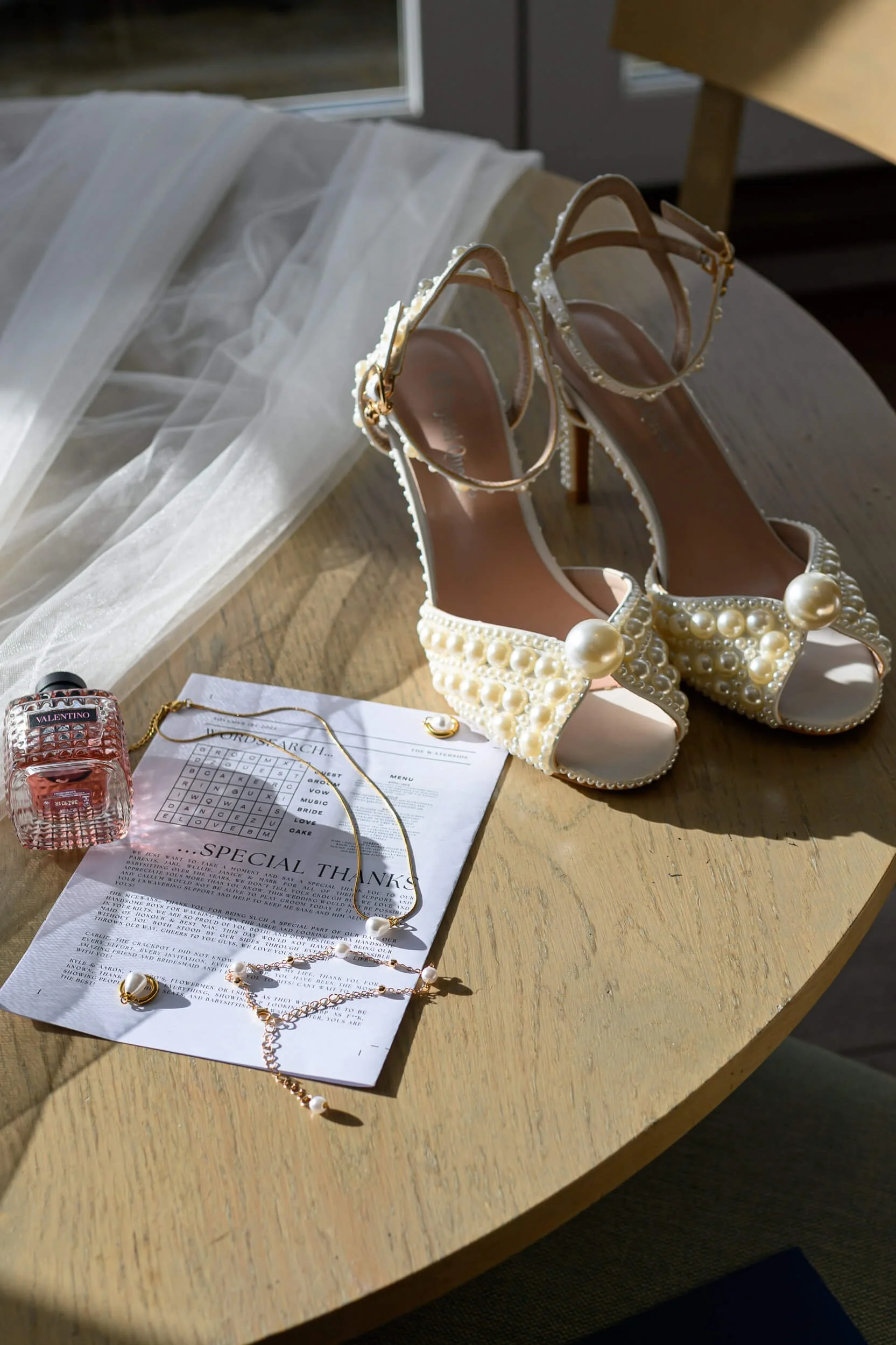 Elegant bridal details captured in soft natural window light at the Waterside Hotel in West Kilbride. Pearl-embellished wedding heels, delicate jewellery, veil, and perfume are beautifully styled on a wooden table, creating a timeless and romantic fl