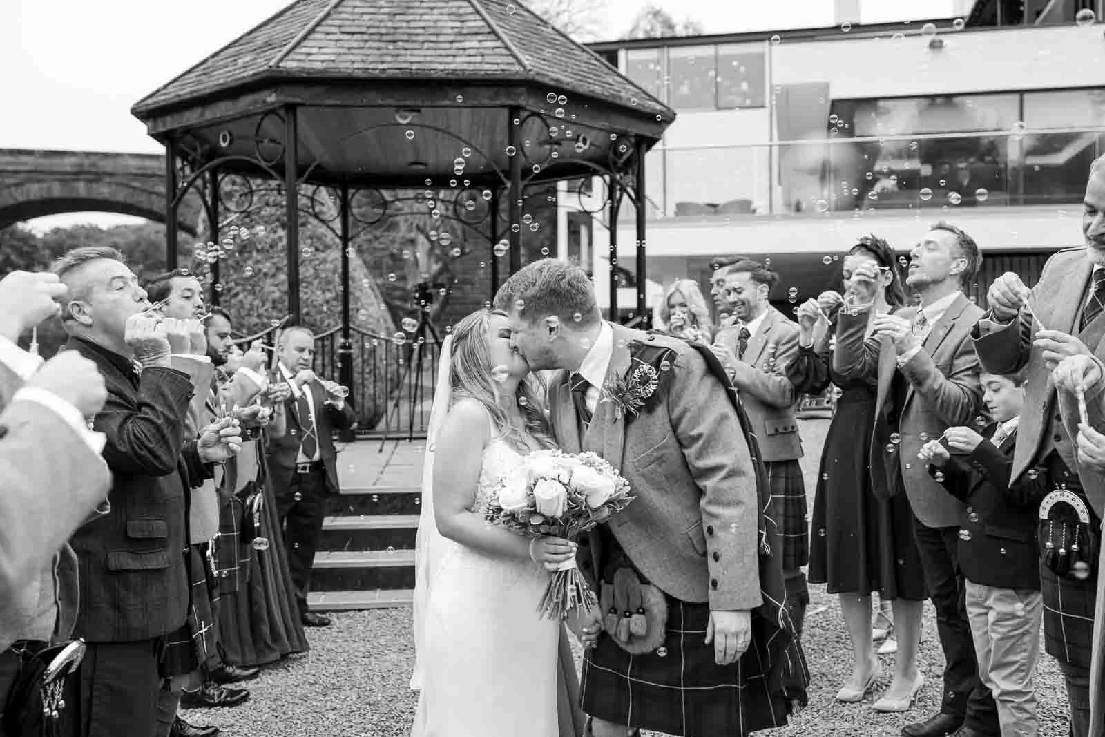 A black and white photo of a bride and groom in a kilt kissing under a shower of bubbles at an outdoor wedding ceremony in Ayr.