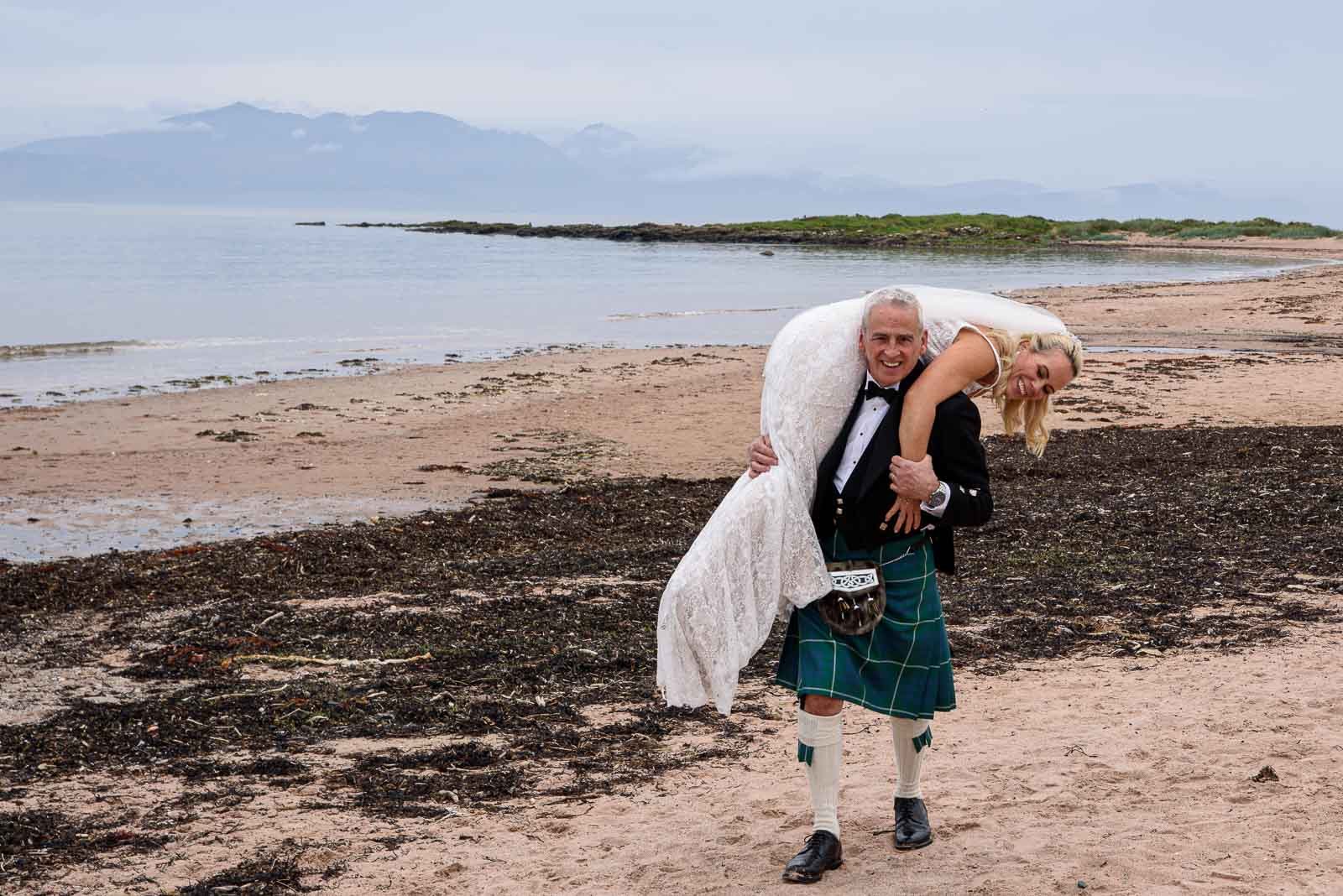 A fun and relaxed wedding moment of a groom lifting his bride on a beach in Ayrshire, Scotland.