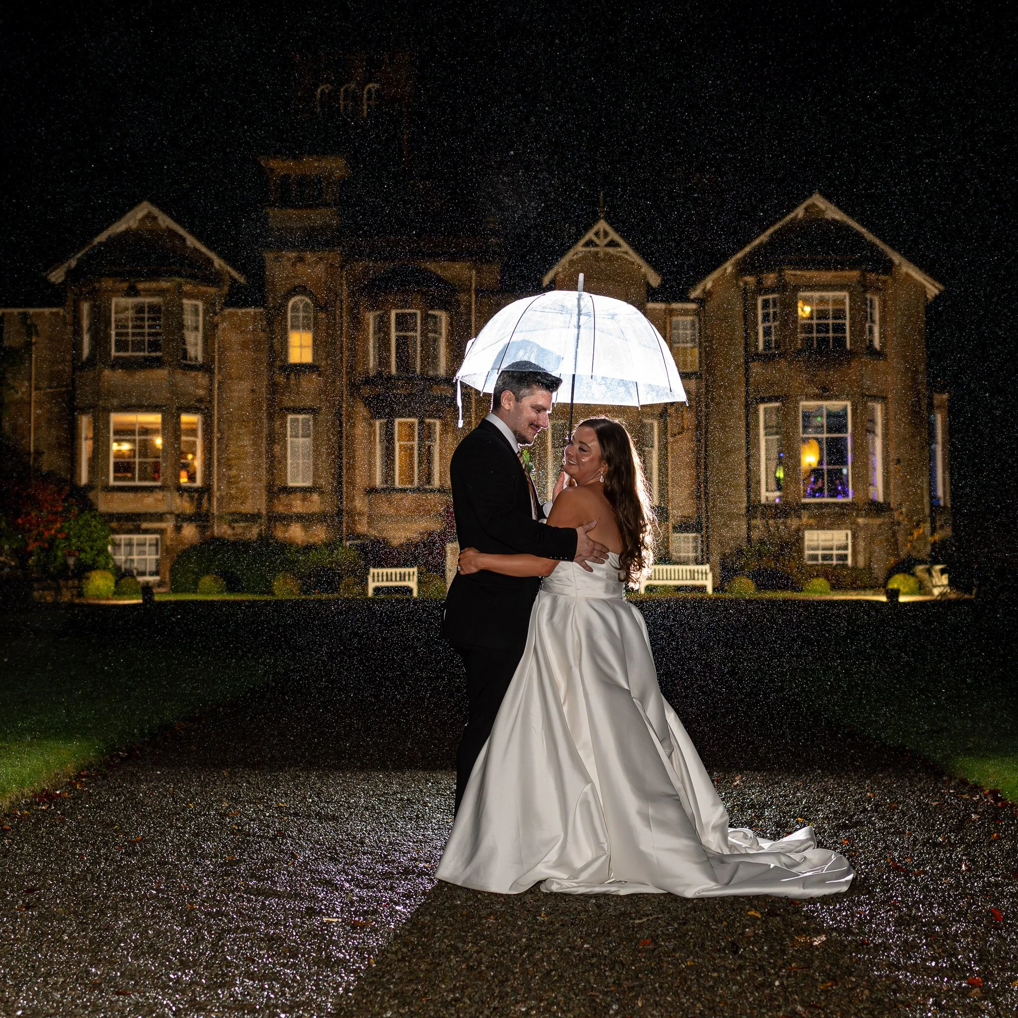 A bride and groom dancing under a transparent umbrella on a rainy night in front of a large, illuminated stone mansion.