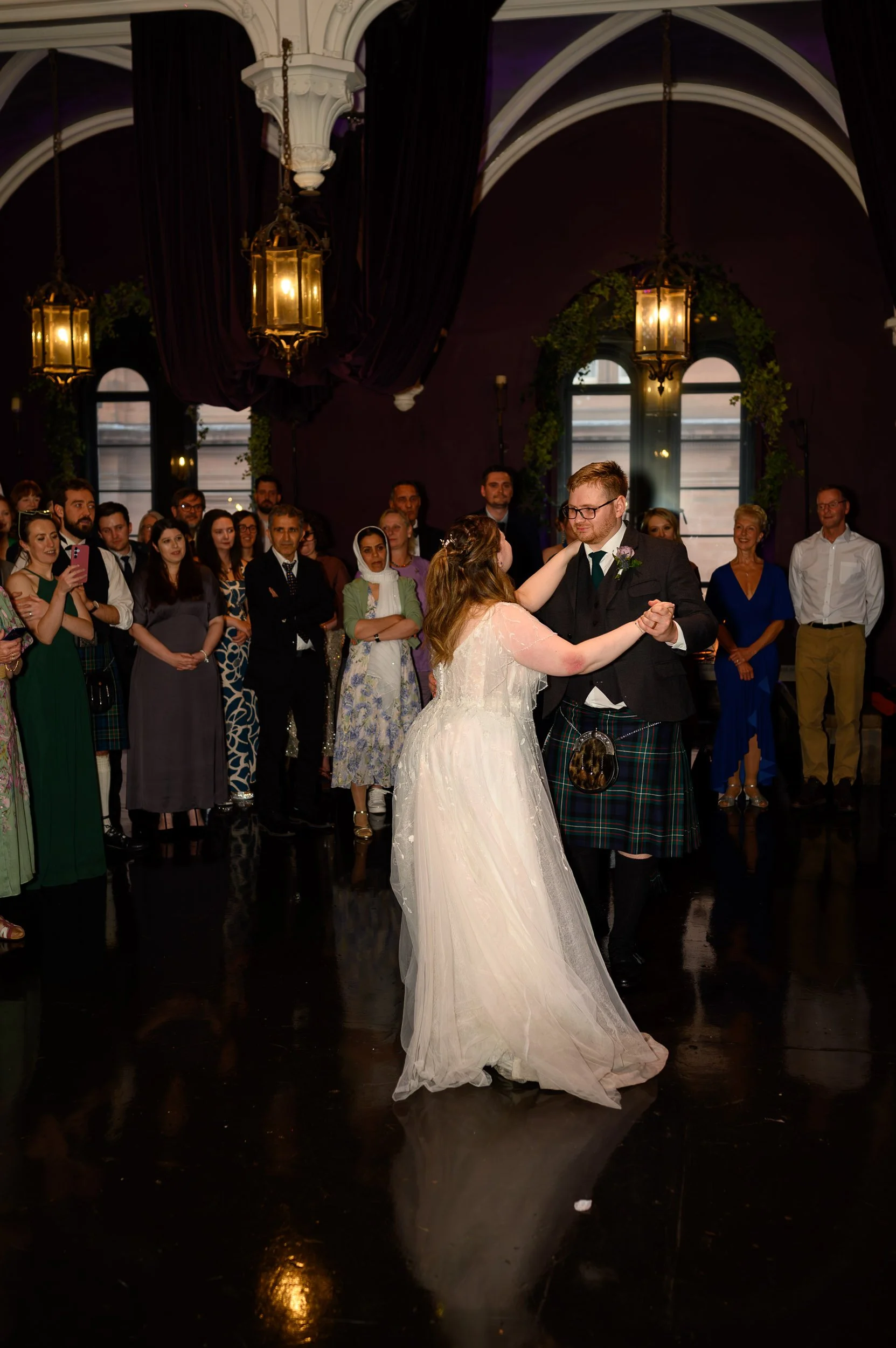 A romantic first dance at The Ghillie Dhu in Edinburgh, captured in the glow of warm lantern light and dramatic dark interiors. The bride and groom share an intimate moment on the dance floor, surrounded by friends and family in the stunning vaulted 