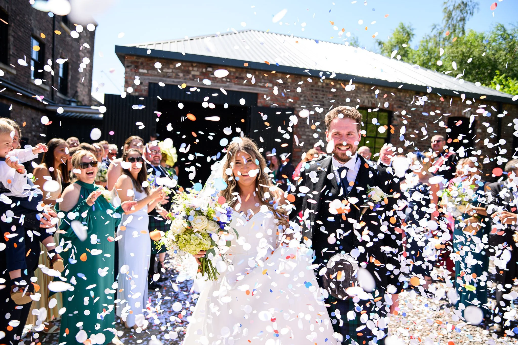 Bride and groom walking through confetti with wedding guests outdoors on a sunny day.