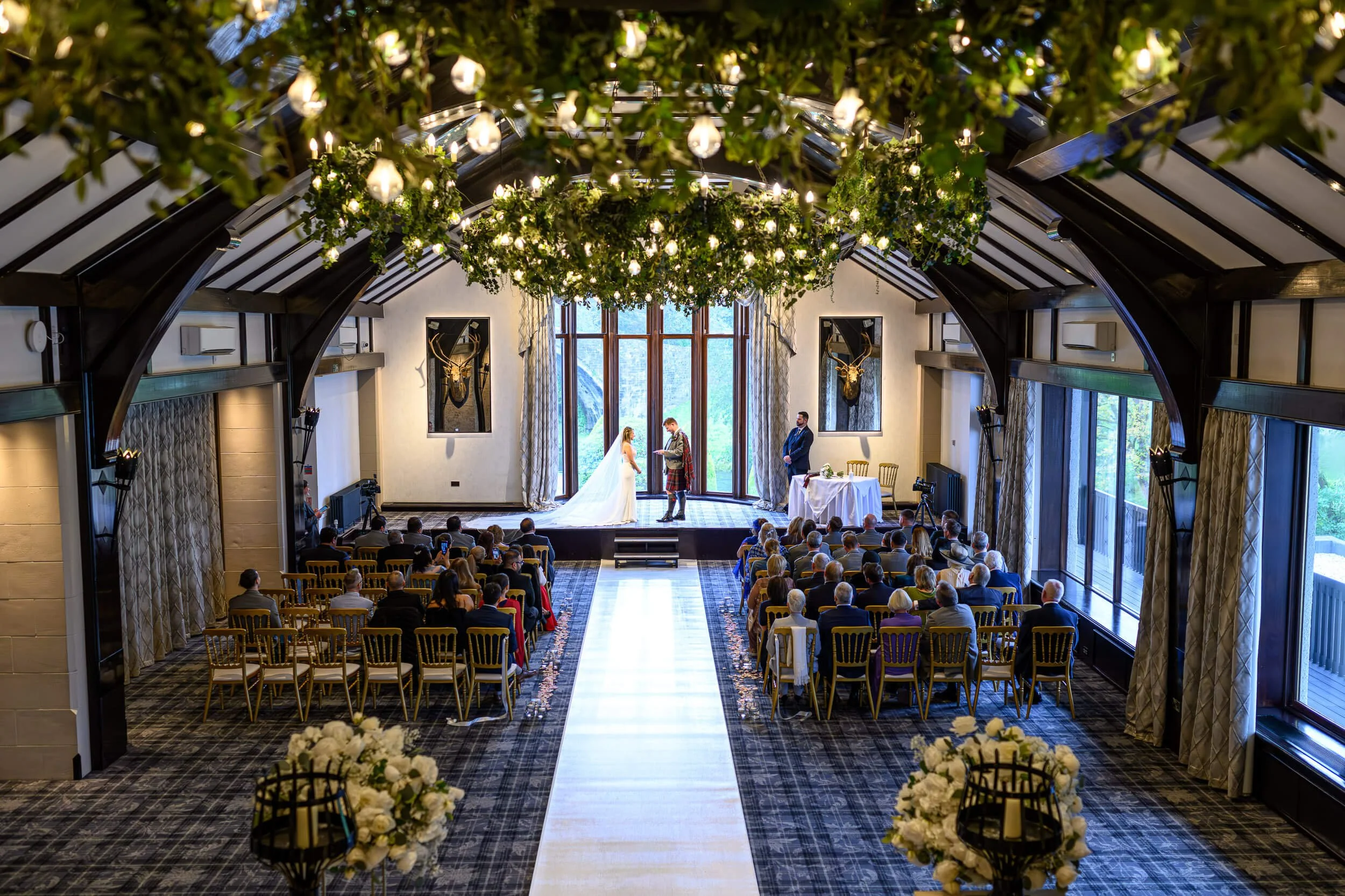 A wide, symmetrical ceremony photograph at Brig O’Doon in Alloway, featuring the bride and groom standing at the altar beneath a stunning greenery installation with hanging warm lights. The aisle leads centrally toward the couple, framed by dark timb