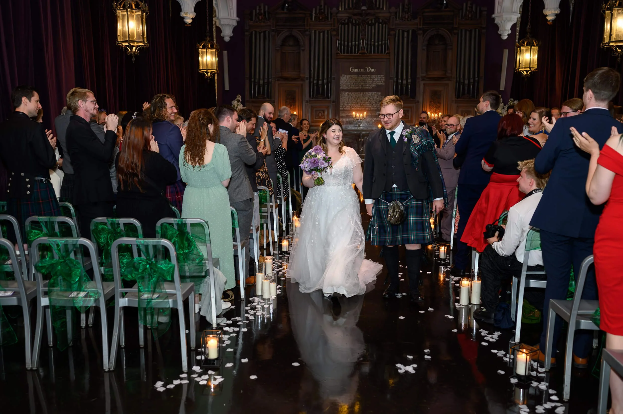 Bride and groom walking hand in hand down the aisle after their candlelit ceremony at Ghillie Dhu in Edinburgh. Guests stand and applaud as the newlyweds celebrate their marriage, surrounded by warm lighting, rose petals and traditional Scottish deta