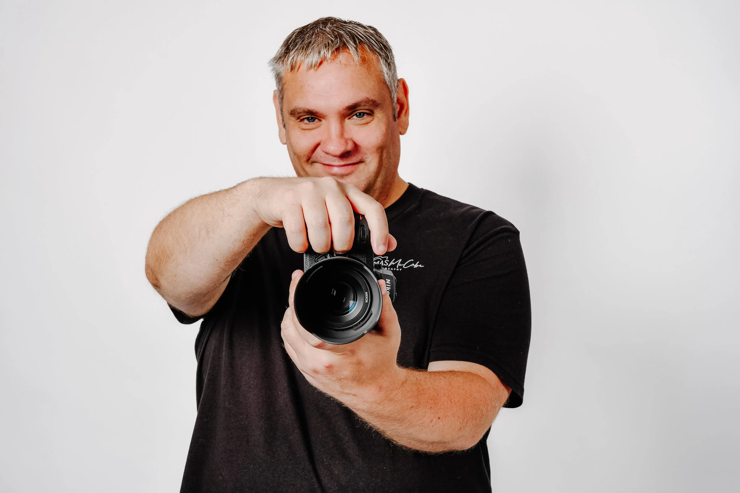 Thomas, a friendly professional photographer in Ayr, smiling warmly while holding a Nikon camera toward the viewer.