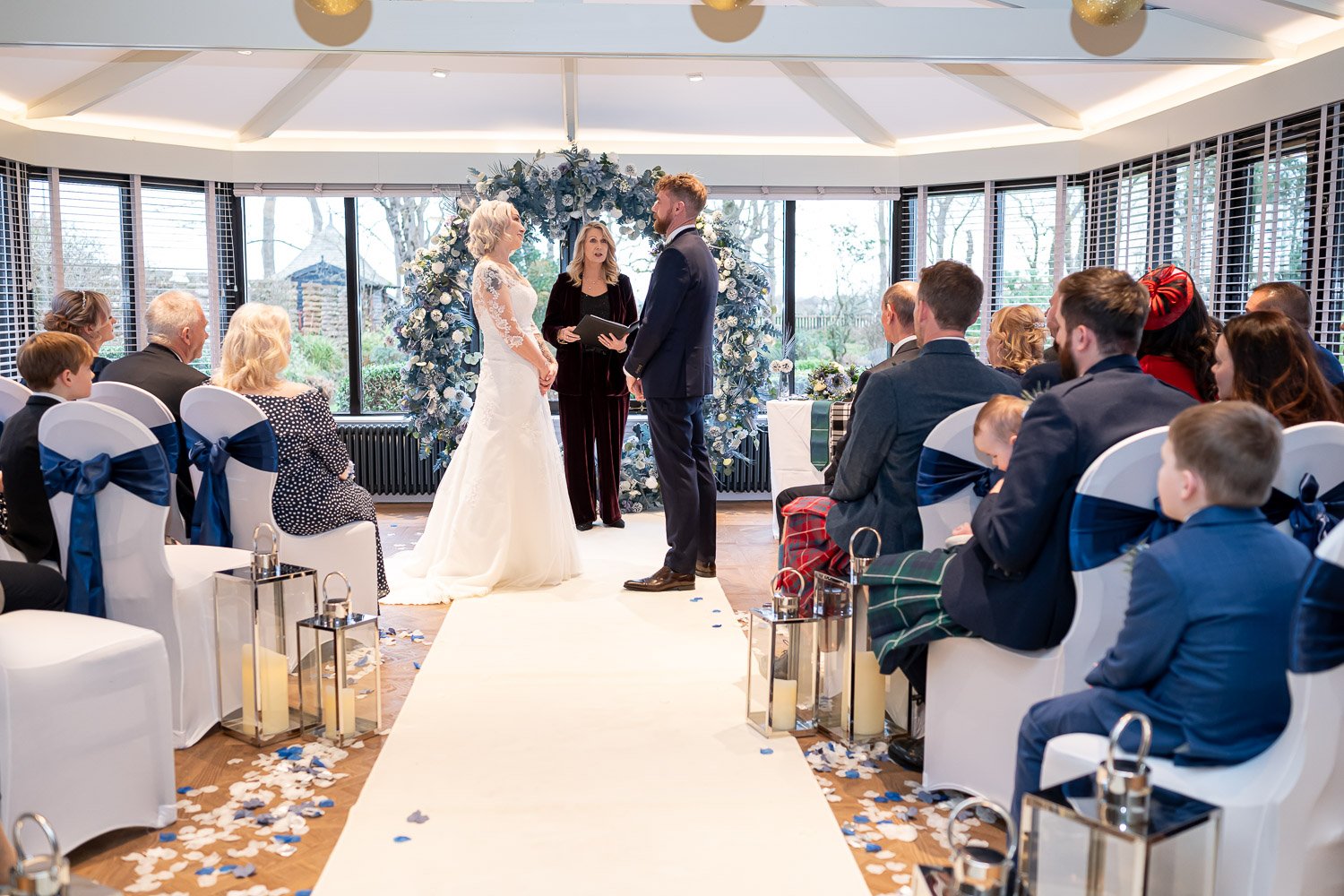 A bright wedding ceremony taking place in the conservatory at Piersland House with a large blue floral arch as a backdrop.