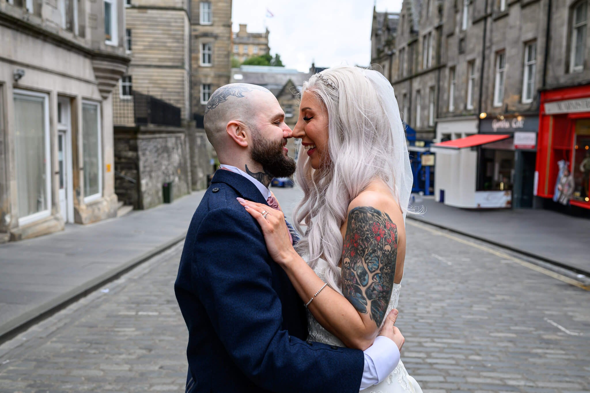 A close-up, intimate portrait of a tattooed bride and groom laughing together on a cobbled street in Edinburgh, surrounded by historic stone buildings.