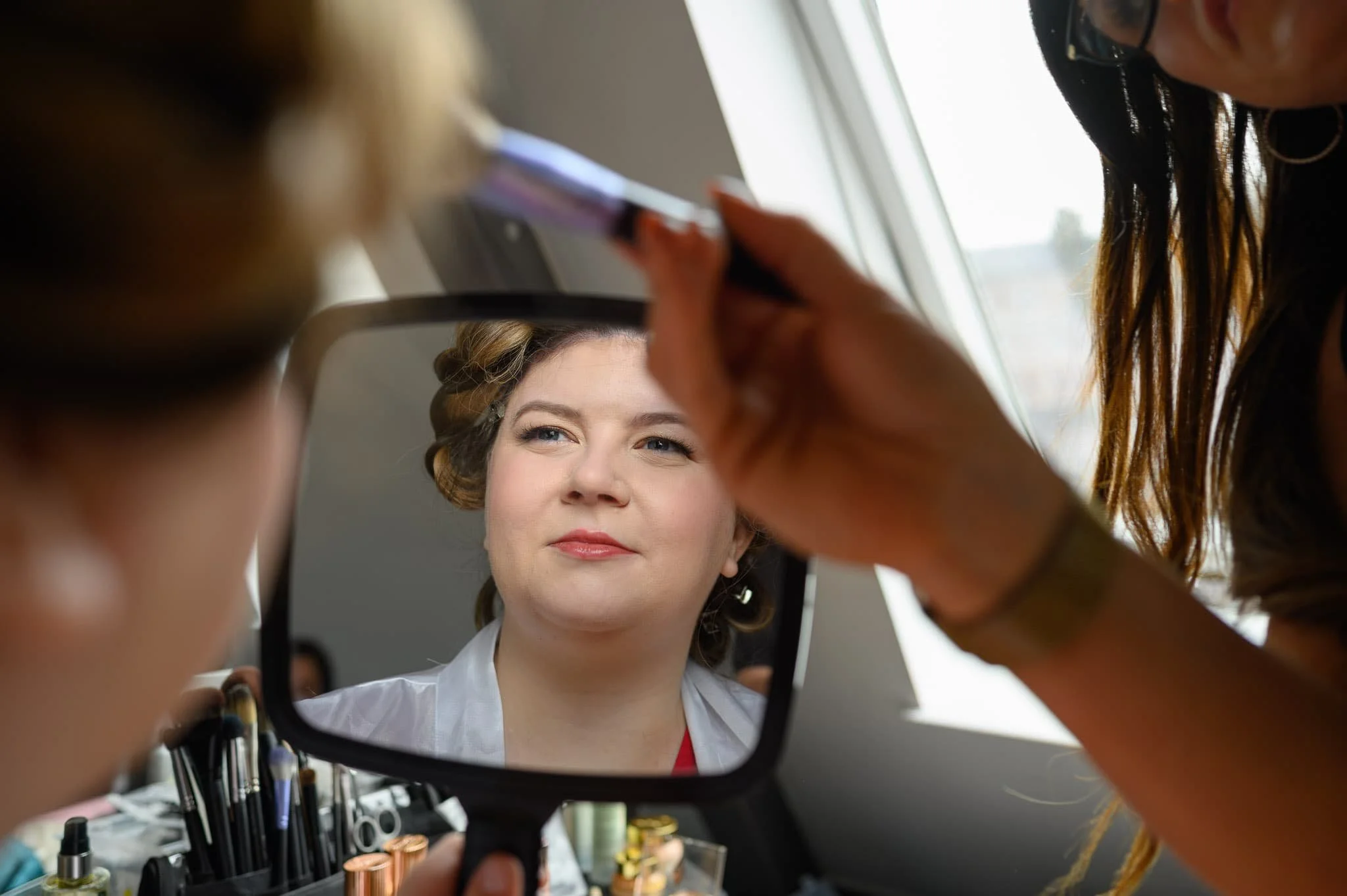 Bride looking at her reflection in a mirror while having her makeup applied during bridal preparations at North Bridge, Edinburgh.
