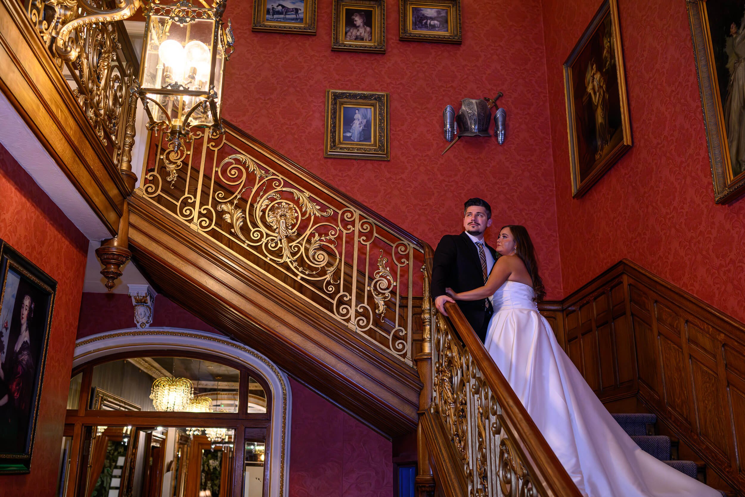 A dramatic wedding portrait of the newlyweds posed on the grand staircase at Auchen Castle Hotel in Moffat. Surrounded by rich red walls, ornate gold detailing, and framed artwork, the bride’s flowing gown cascades down the staircase as the couple sh