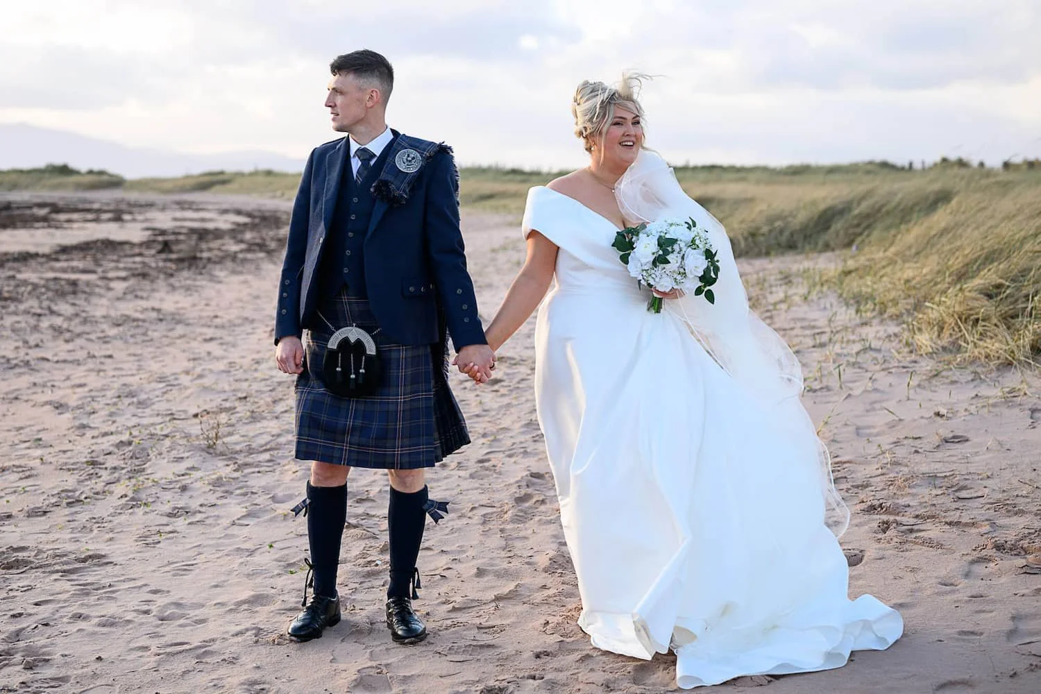 Bride and groom walking hand in hand along the beach at The Waterside Hotel in West Kilbride, Ayrshire. With the coastline stretching behind them and the breeze catching the bride’s veil, this relaxed seaside portrait captures the natural, romantic a