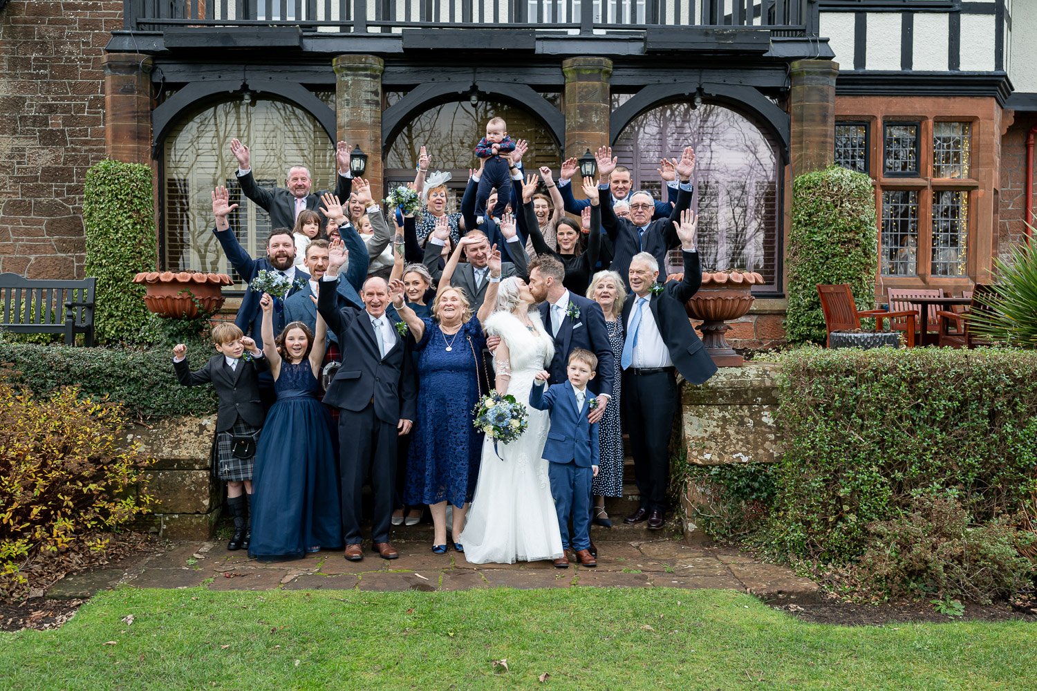 A large wedding party cheering with their hands in the air on the stone steps outside Piersland House Hotel.