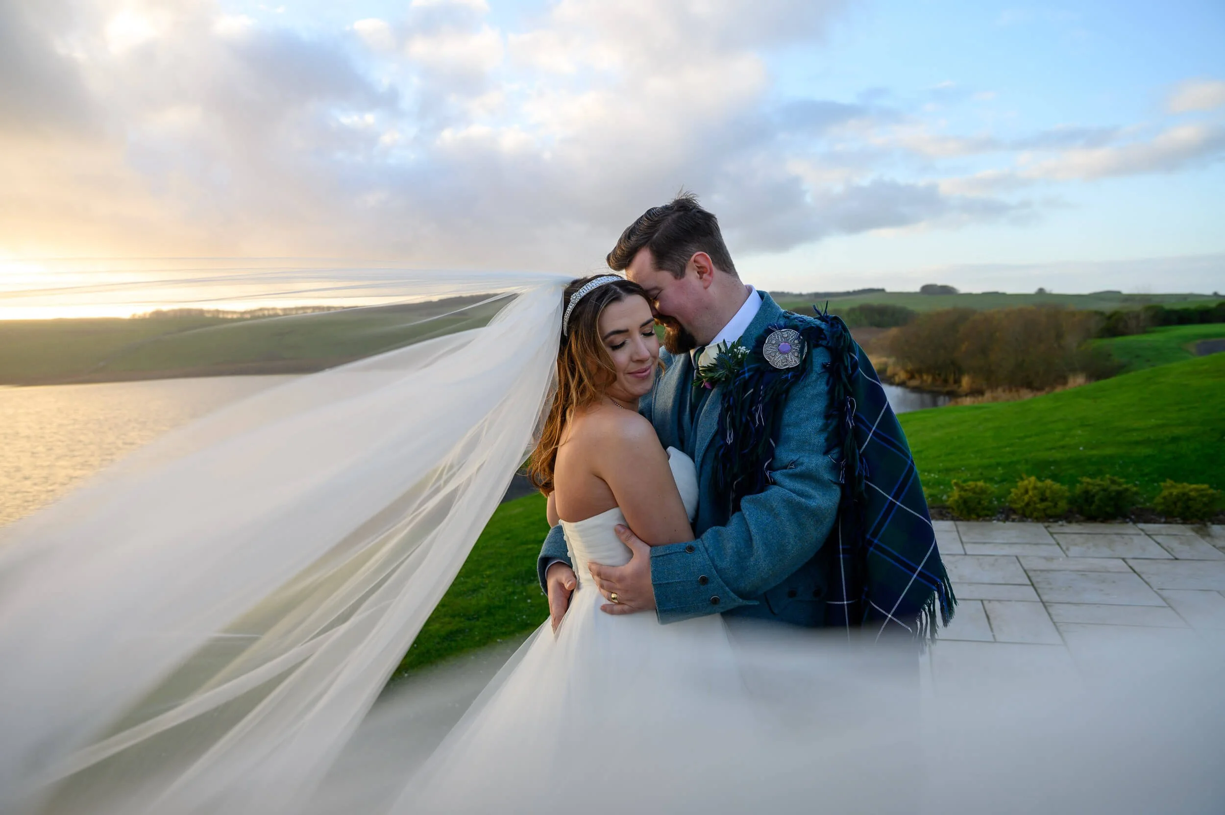 Romantic sunset wedding portrait at Lochside Hotel, New Cumnock. The bride and groom embrace as the evening light glows over the water, with the bride’s veil flowing beautifully in the wind. A natural, emotional moment captured overlooking the Ayrshi